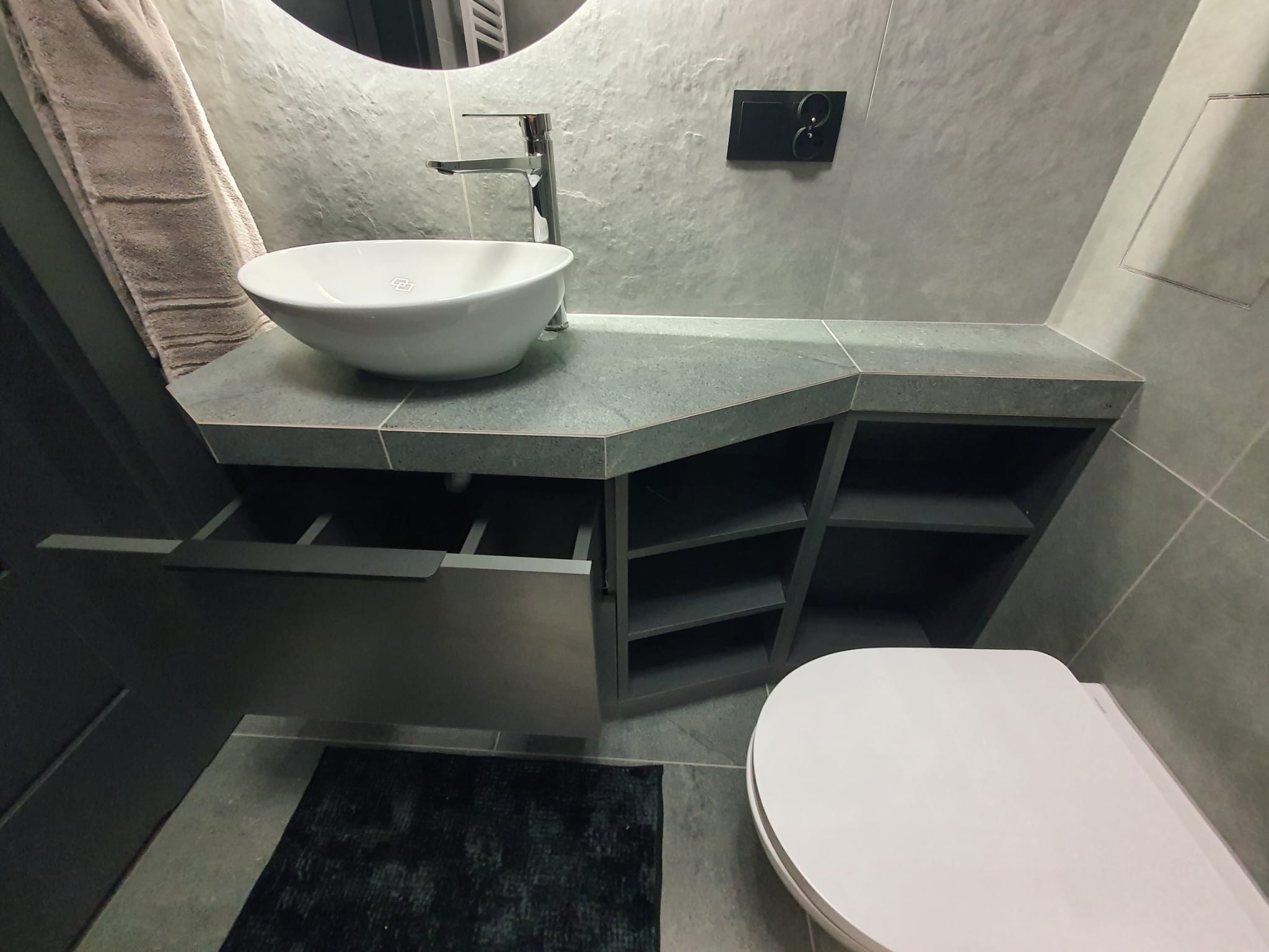 Nowoczesna łazienka z umywalką nablatową, szarą zabudową meblową z otwartymi półkami i czarnym dywanikiem. Minimalistyczny design, płytki imitujące kamień.