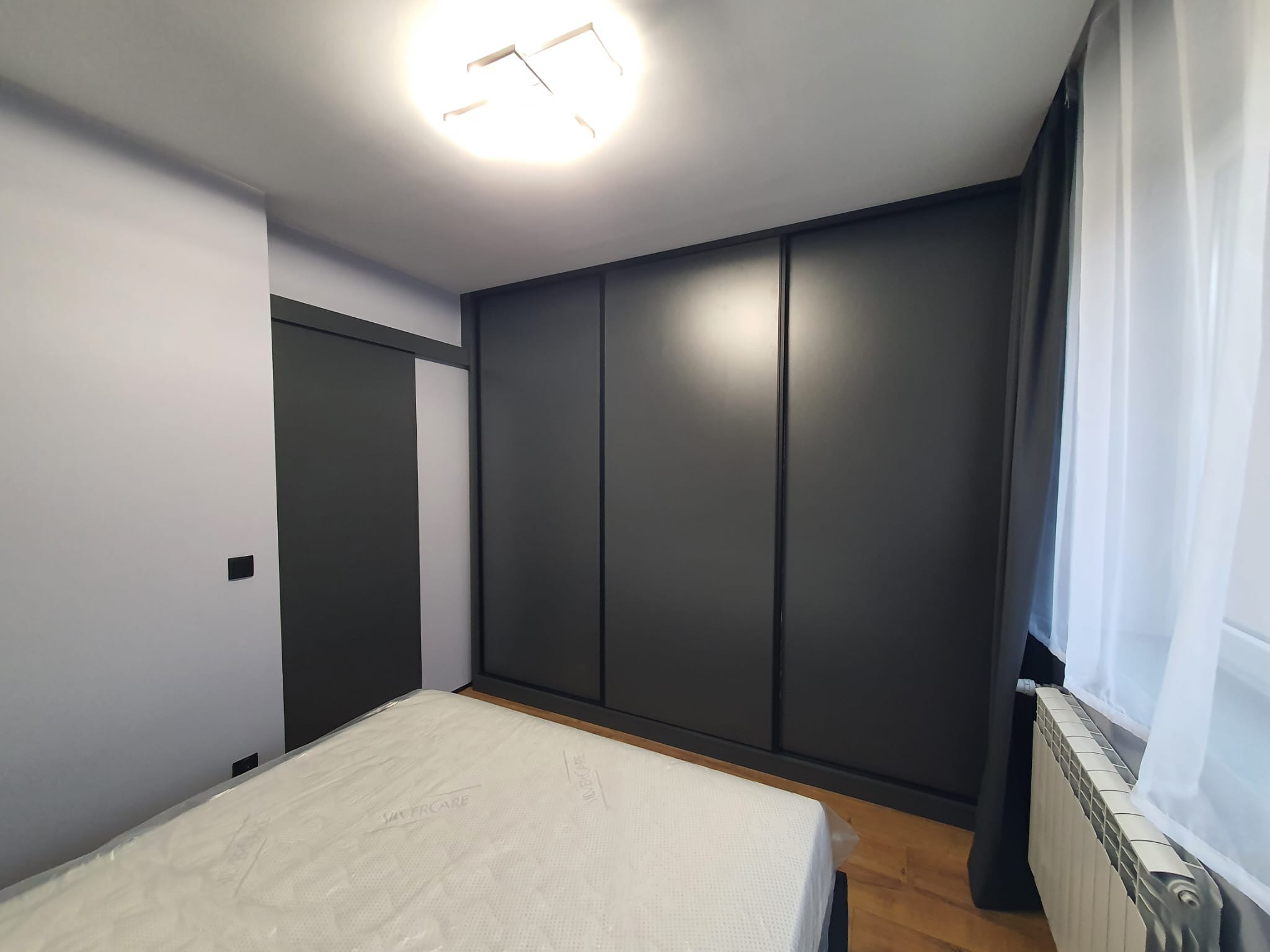 Nowoczesna szafa przesuwna w ciemnym kolorze, idealnie wkomponowana w jasne wnętrze sypialni. Minimalistyczny design, gładkie fronty, subtelne oświetlenie sufitowe.