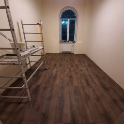 Sławomir Oryga - Ukończone układanie paneli podłogowych w pokoju z oknem w kształcie łuku. Widoczny drabina malarska z narzędziami i farbą. Ciepłe oświetlenie sufitowe.