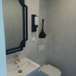 Fachmann - Wykończona łazienka w Nowogardzie: biała umywalka z złotą baterią, lustro w czarnej ramie, toaleta i dekoracyjne płytki. Minimalistyczny design z akcentem elegancji.