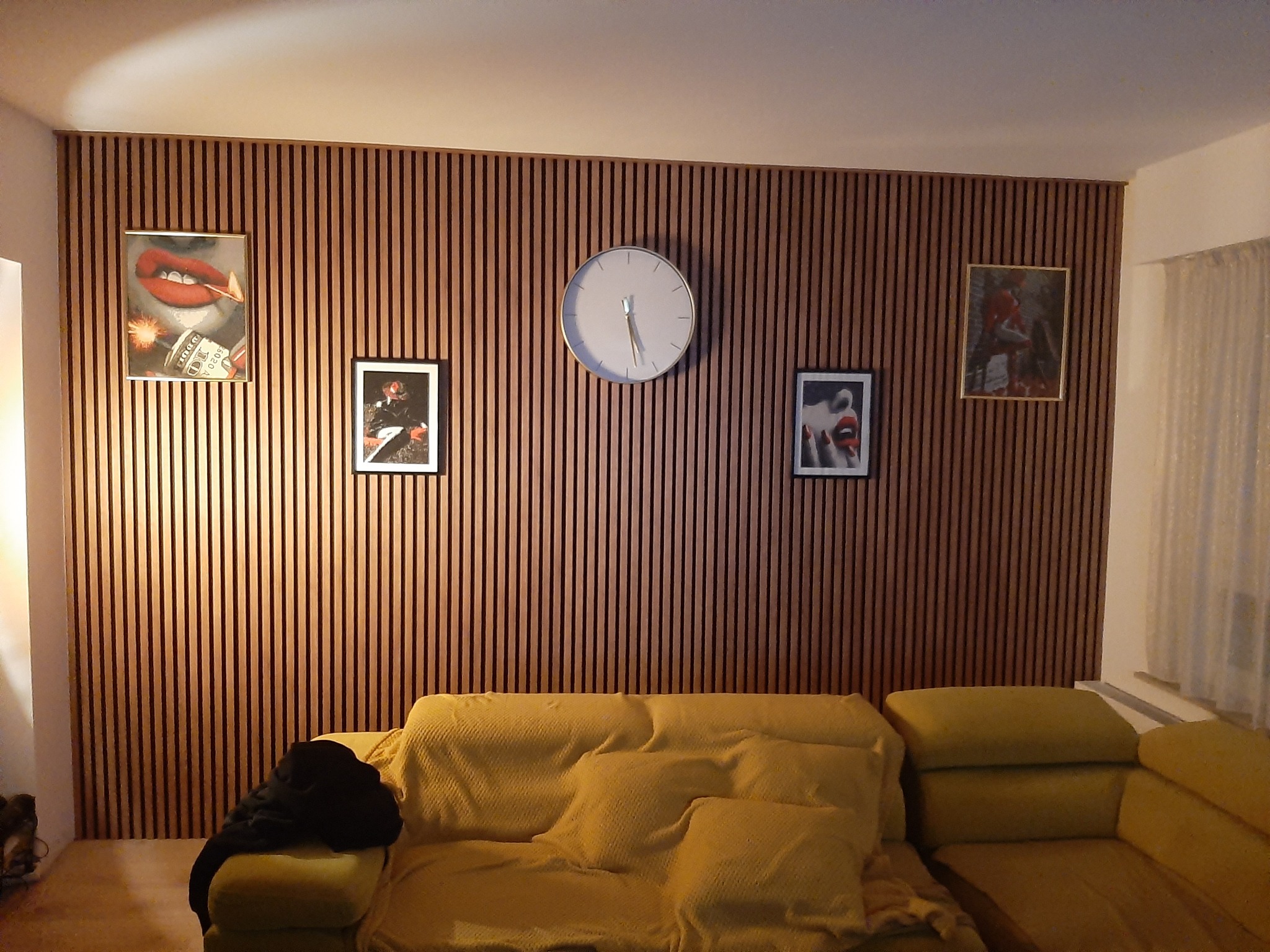 Ściana z drewnianych paneli lamelowych w salonie, z wiszącymi obrazami i zegarem. Żółta kanapa stoi pod ścianą. Widoczne elementy dekoracyjne.