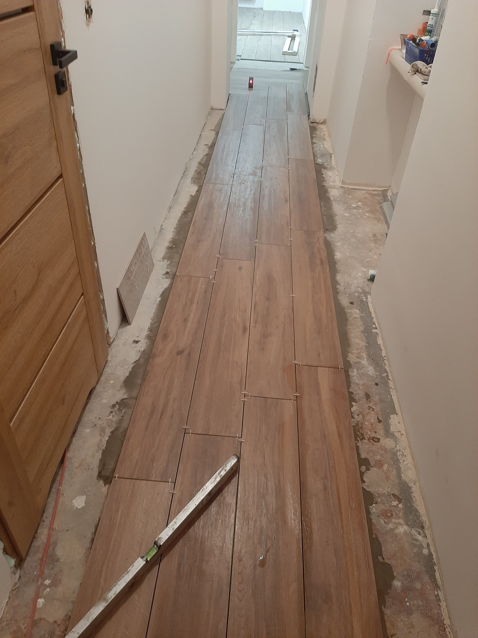Układanie podłogi z płytek imitujących drewno w korytarzu. Widoczny poziomica i krzyżyki dystansowe. Prace w toku, surowy stan ścian.
