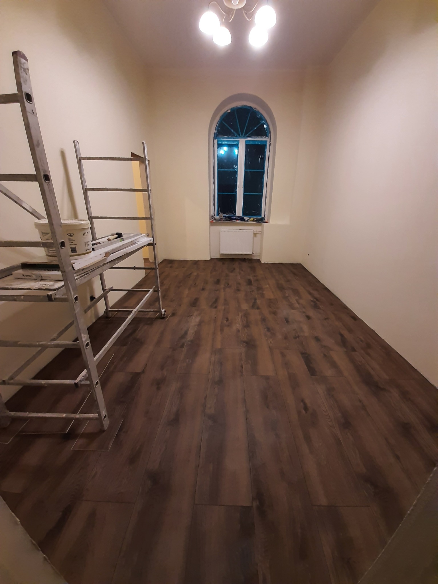 Ukończone układanie paneli podłogowych w pokoju z oknem w kształcie łuku. Widoczny drabina malarska z narzędziami i farbą. Ciepłe oświetlenie sufitowe.