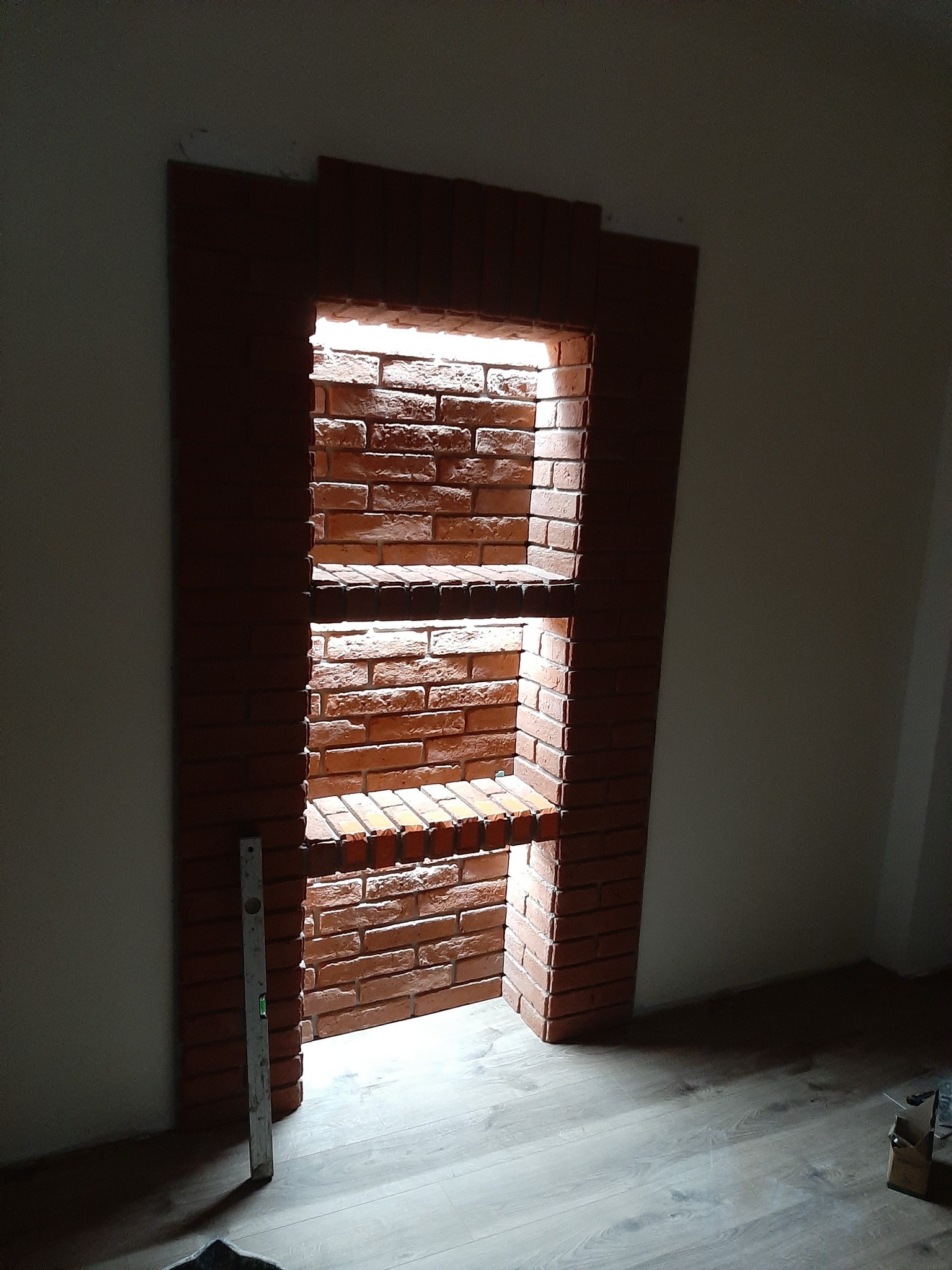 Narożna wnęka z cegły klinkierowej z półkami, oświetlona od góry, na tle jasnej ściany i podłogi. Widoczny poziomica. Dekoracyjne wykończenie wnętrza.