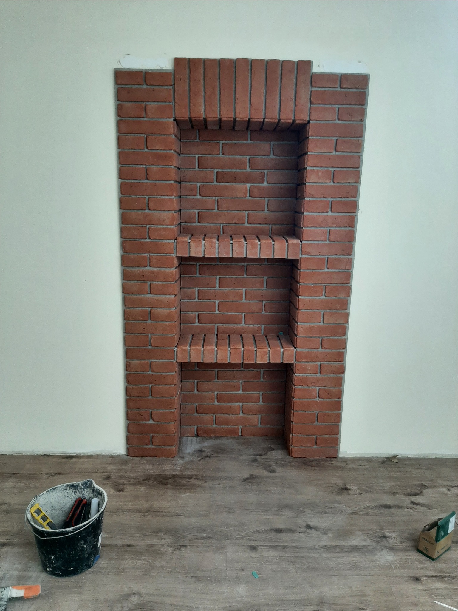 Narożna półka z czerwonej cegły na ścianie, z widocznym wiadrem i narzędziami na podłodze. Wykończenie wnętrza w trakcie realizacji.