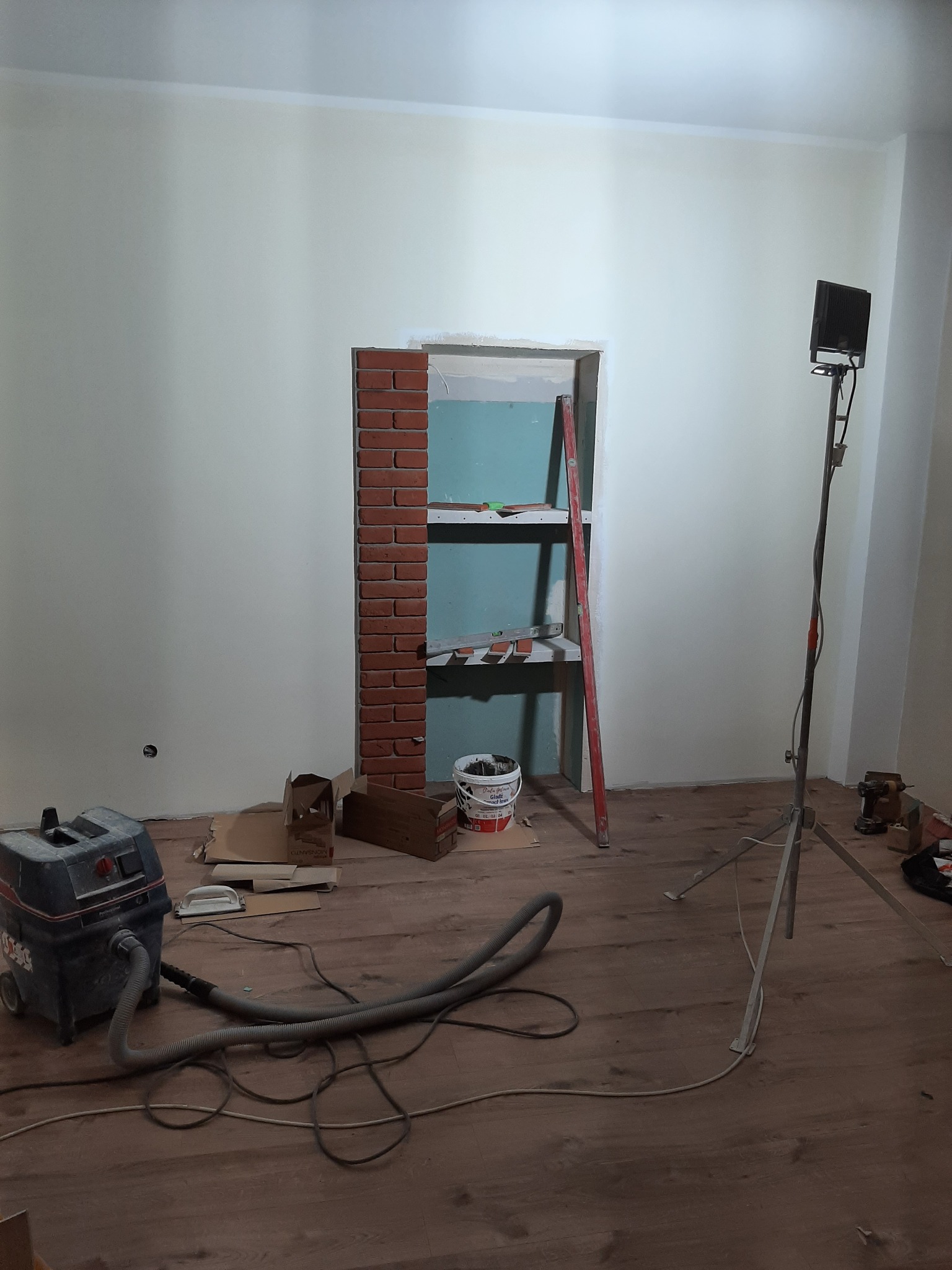 Wnętrze w trakcie prac wykończeniowych: ściana z wnęką obłożoną cegłą, widoczne narzędzia, drabina i odkurzacz przemysłowy na drewnianej podłodze.