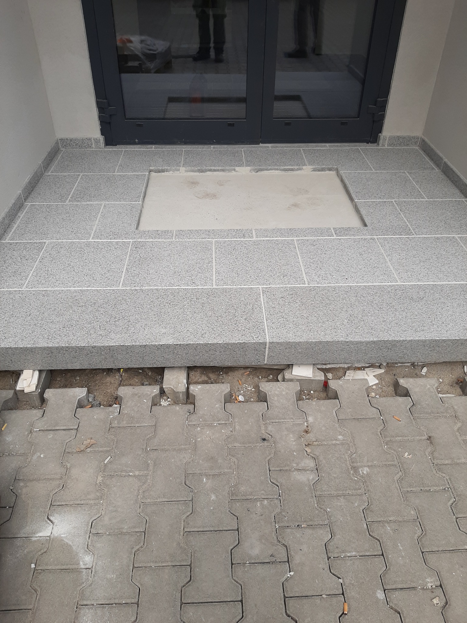 Szare płytki na balkonie w trakcie układania, widoczny fragment podłoża z kostki brukowej. Częściowo wykończona powierzchnia przy szarych drzwiach balkonowych.