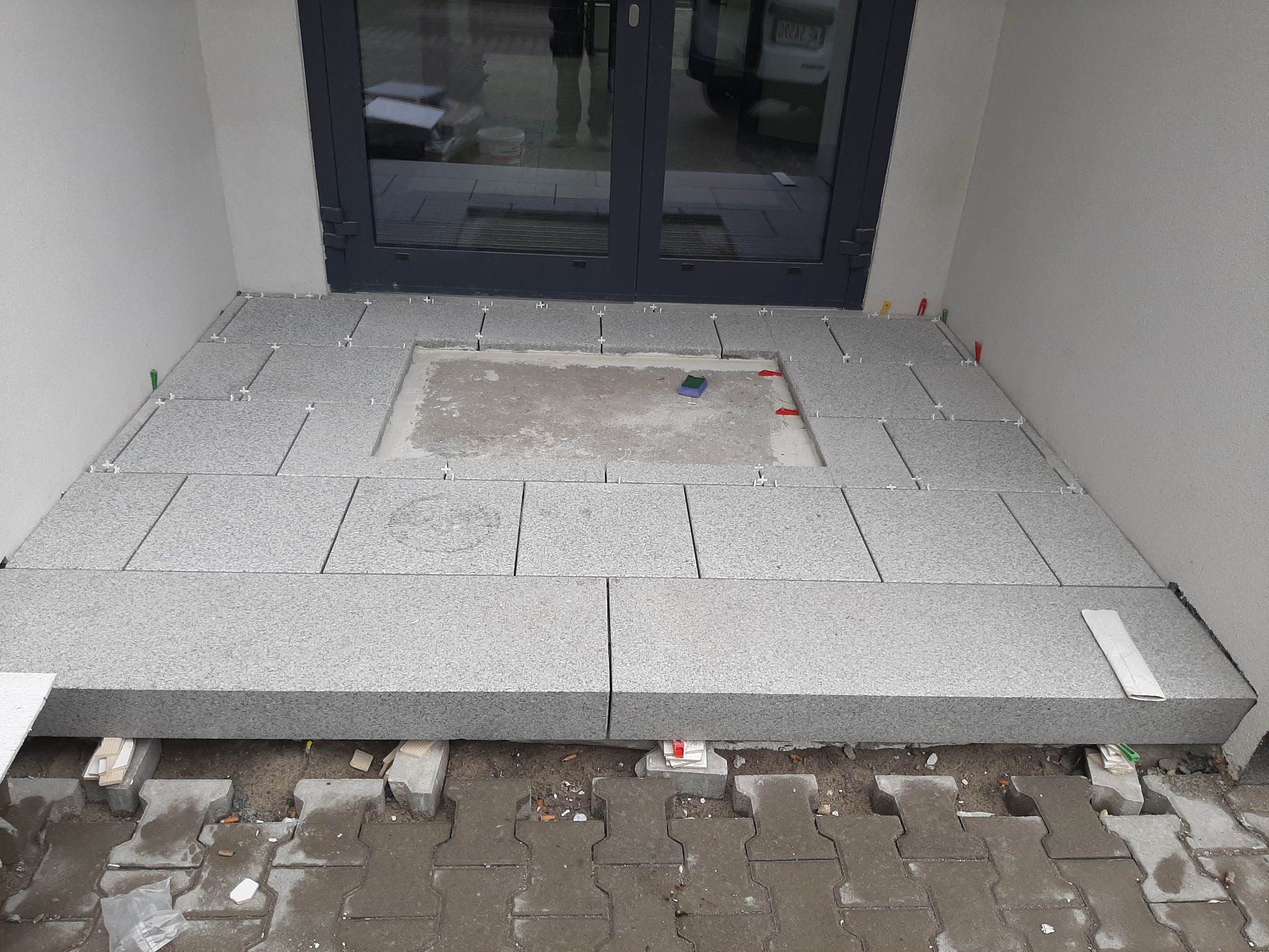 Montaż szarych płyt na balkonie, widoczne podkładki poziomujące i fragment starej nawierzchni. Prace w toku, płytki ułożone częściowo, widoczne szczeliny dylatacyjne.