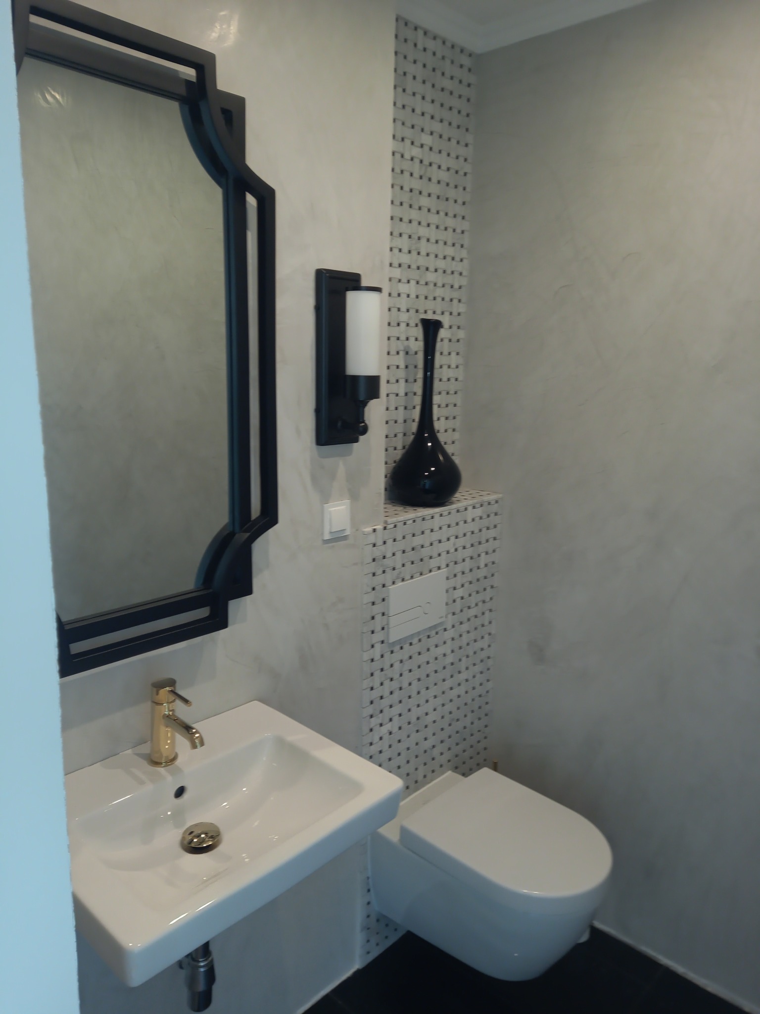 Wykończona łazienka w Nowogardzie: biała umywalka z złotą baterią, lustro w czarnej ramie, toaleta i dekoracyjne płytki. Minimalistyczny design z akcentem elegancji.