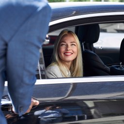 PACZOS PIOTR AUTO PASJA - Uśmiechnięta blondynka w czarnym samochodzie, otwierana przez mężczyznę w niebieskim garniturze. Luksusowy transport.