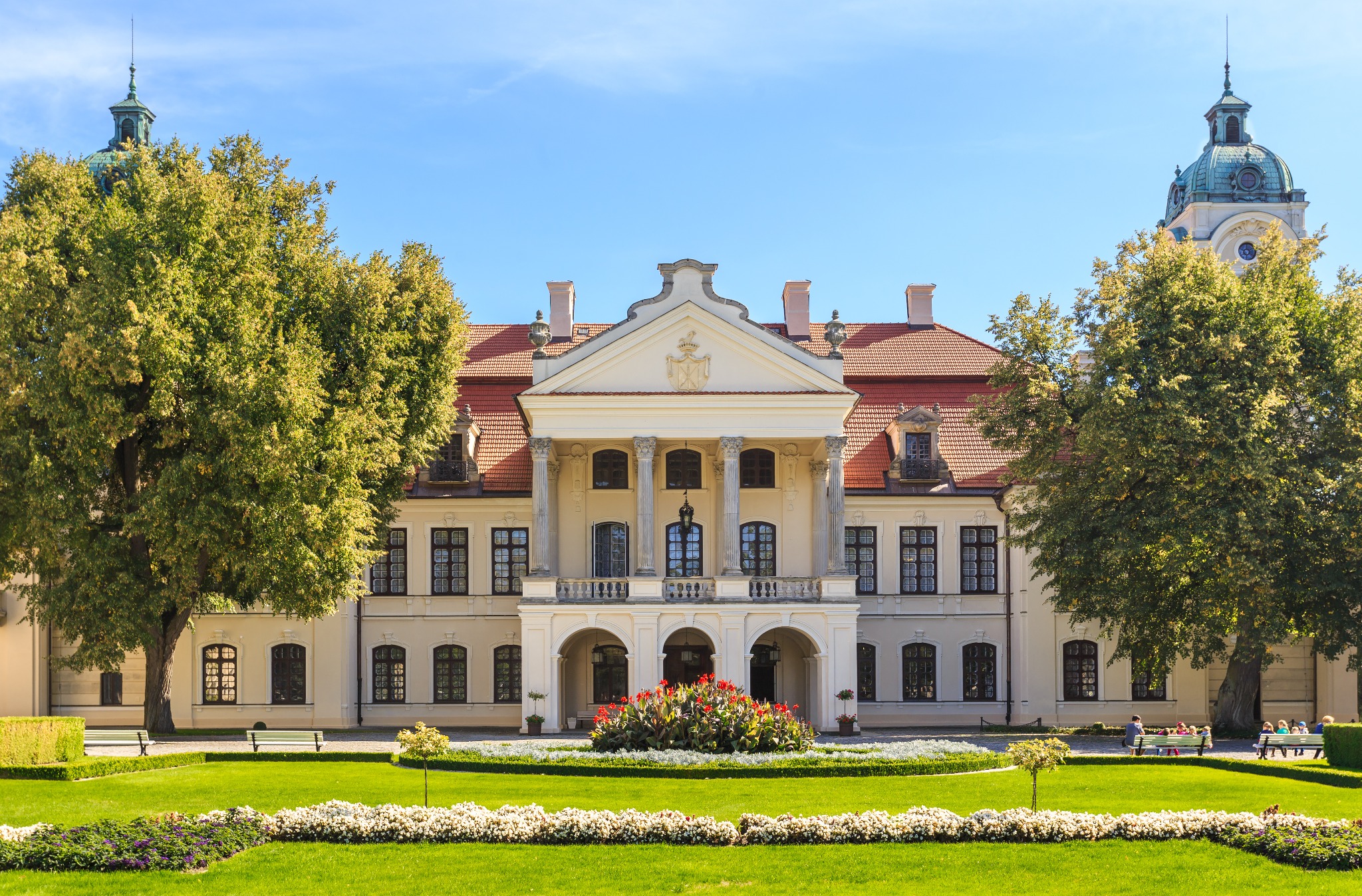 Barokowy pałac z kolumnami i wieżą zegarową, otoczony zielonym trawnikiem i rabatami kwiatowymi w słoneczny dzień. Widoczne ławki i spacerujący ludzie.
