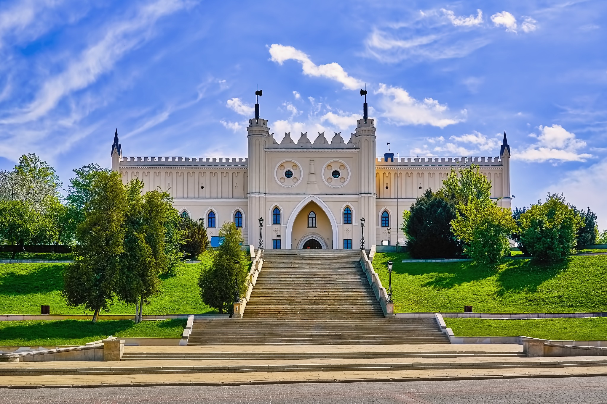 Zamek w Lublinie w słoneczny dzień. Widok z dołu na schody prowadzące do zamku, otoczonego zielenią. Błękitne niebo z delikatnymi chmurami w tle.