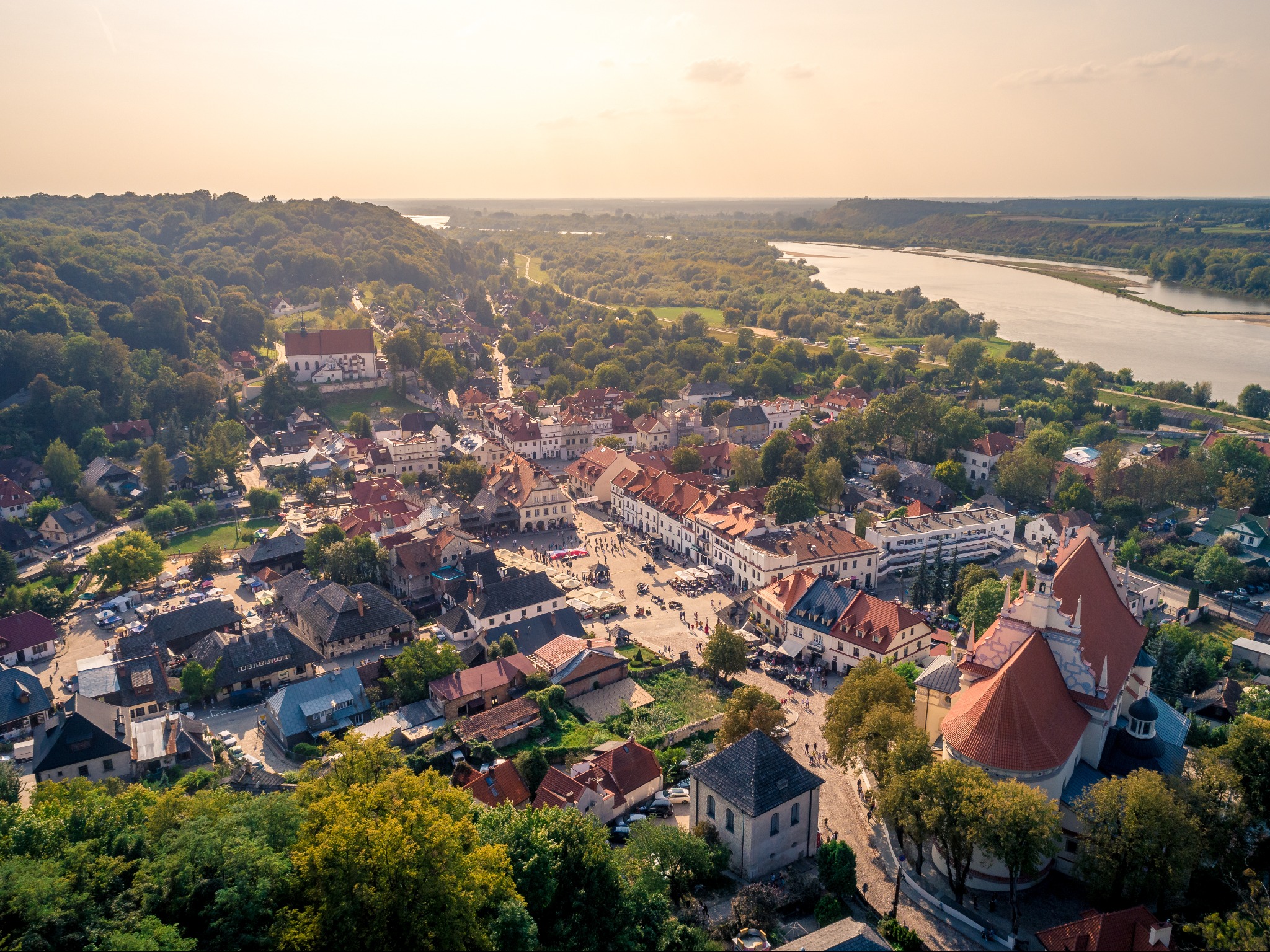 Panoramiczny widok z lotu ptaka na Kazimierz Dolny: rynek z ludźmi, czerwone dachy budynków, zieleń drzew i rzeka w tle, ciepłe światło zachodzącego słońca.