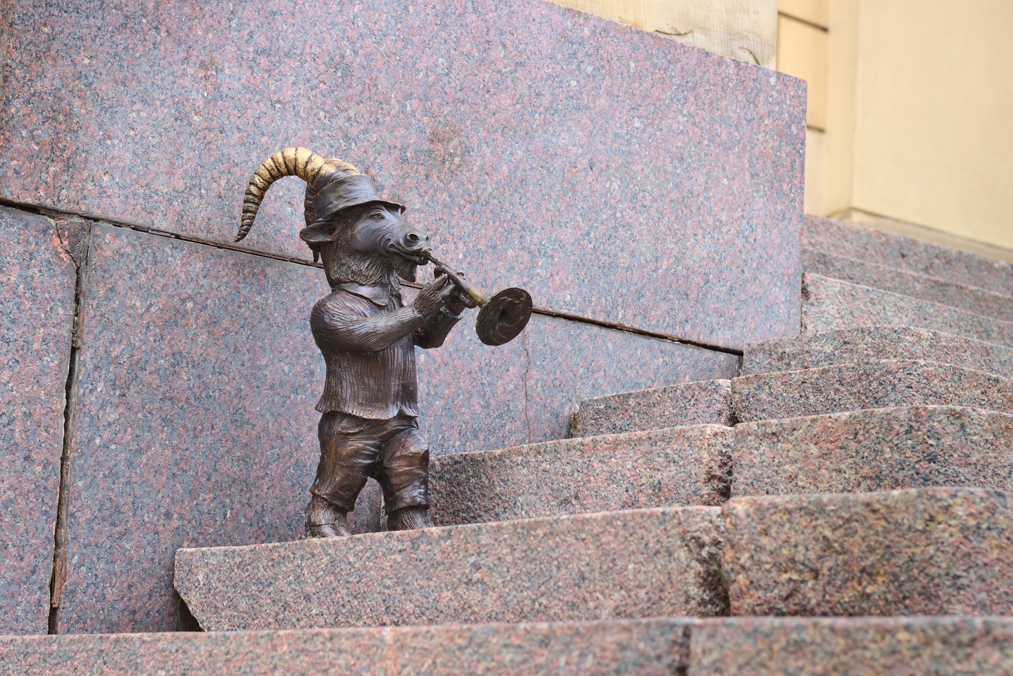 Brązowa rzeźba kozła grającego na trąbce, umieszczona na granitowych schodach. Kozioł ubrany w kapelusz i marynarkę. Nietypowa dekoracja miejska.