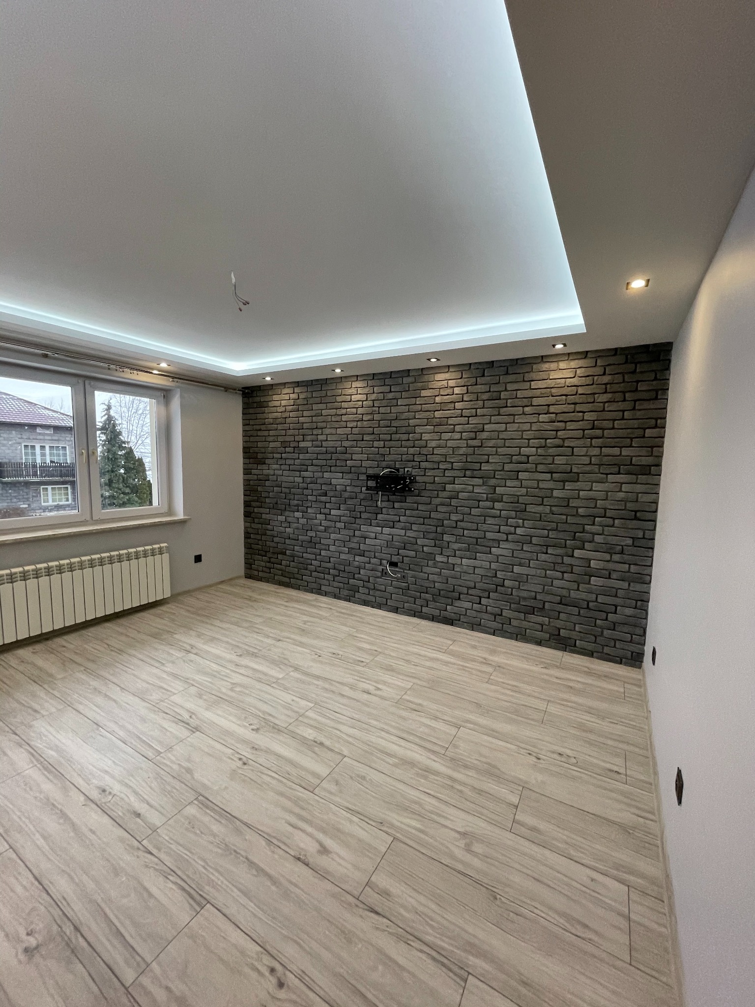 Pokój po remoncie: drewnopodobna podłoga, ściana z szarej cegły, podwieszany sufit z oświetleniem LED. Widok na okno i grzejnik. Minimalistyczny design.
