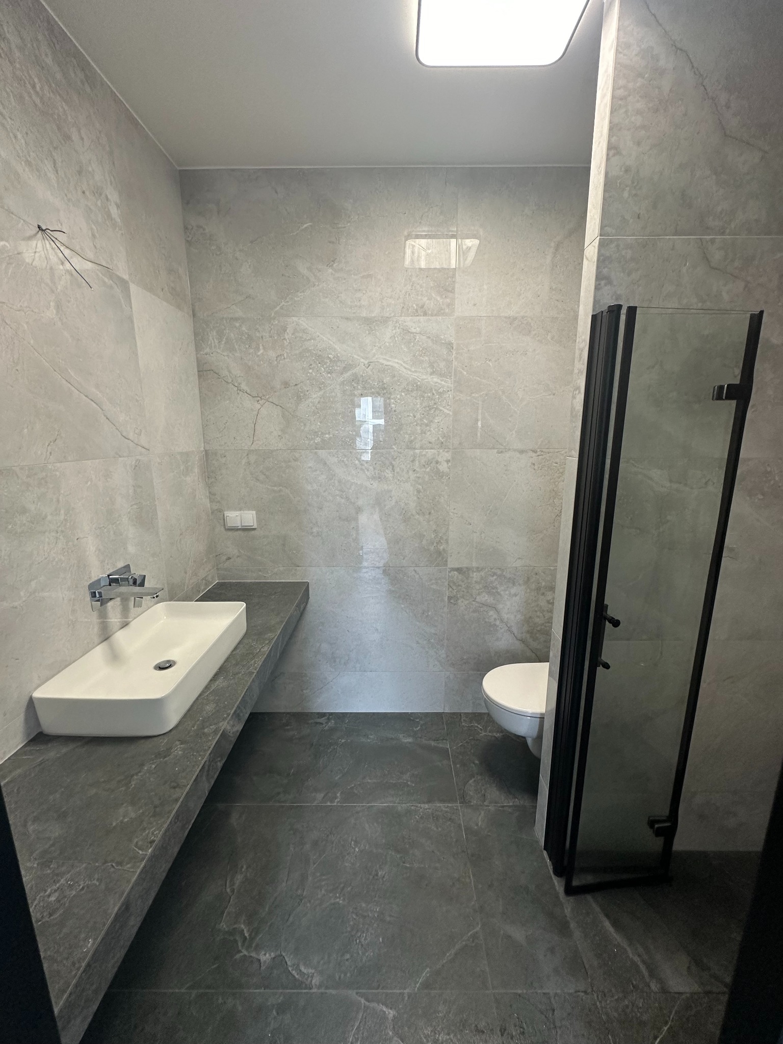 Nowoczesna łazienka w szarościach: marmurowe płytki, prostokątna umywalka na blacie, kabina prysznicowa z czarnymi ramami i wisząca toaleta. Minimalistyczny design.