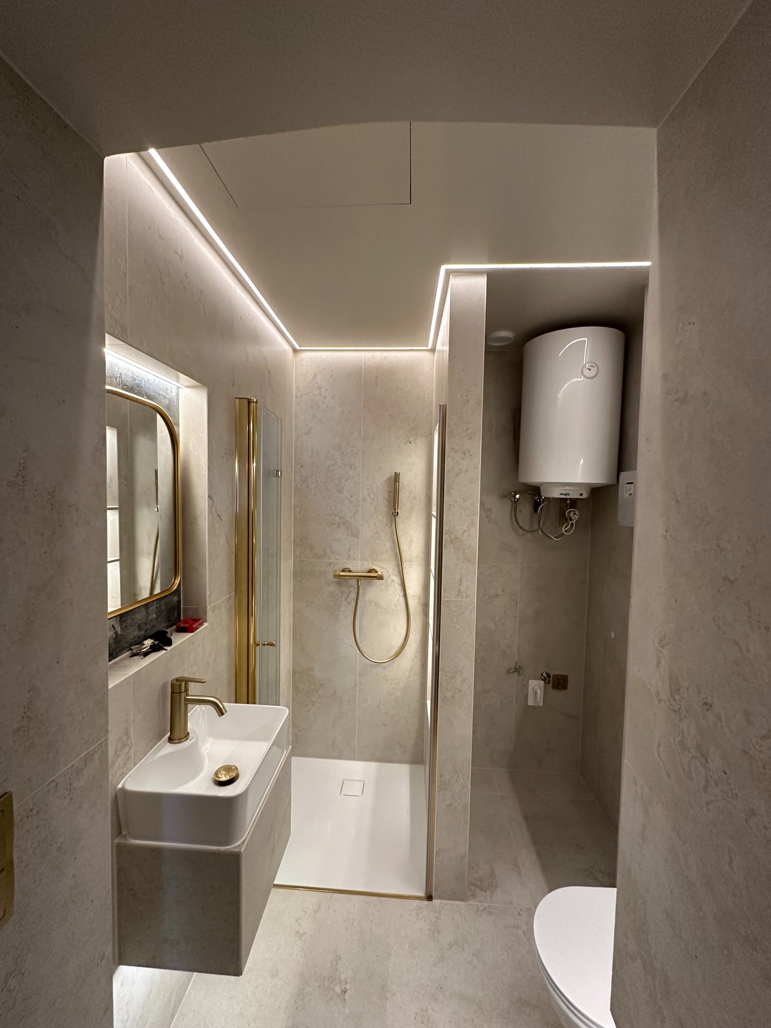 Wykończona łazienka w beżowych płytkach z kabiną prysznicową, umywalką nablatową i podświetleniem LED. Widoczny bojler i złote akcenty.