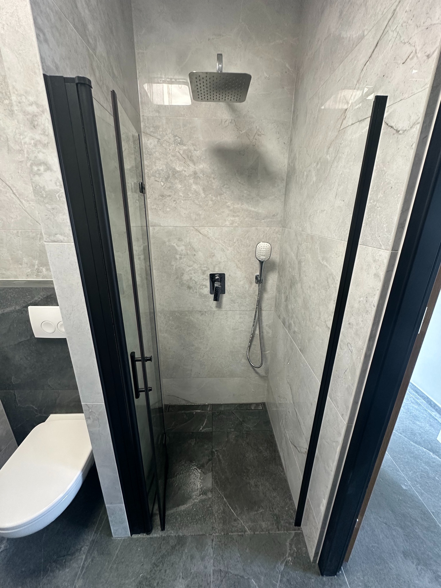Nowoczesna kabina prysznicowa z czarnymi ramami i deszczownicą, wykończona płytkami imitującymi marmur. Widoczna toaleta w tle, szara podłoga.