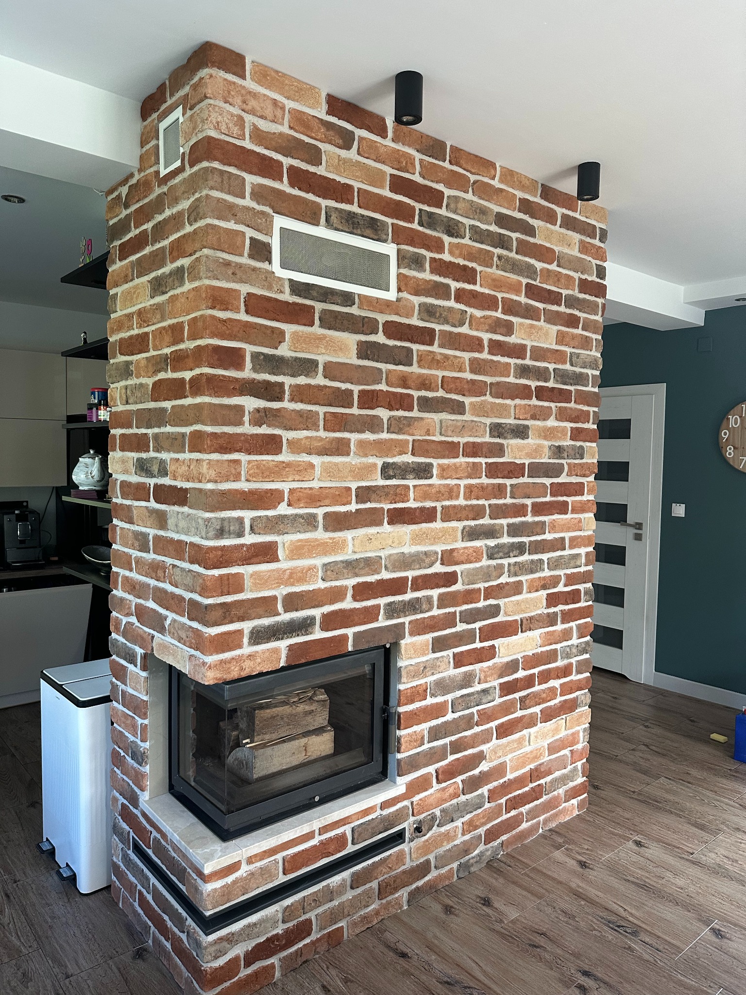 Murowany kominek z cegły klinkierowej w salonie, z widocznym paleniskiem i drewnem w środku. W tle kuchnia i drzwi. Podłoga wyłożona panelami imitującymi drewno.