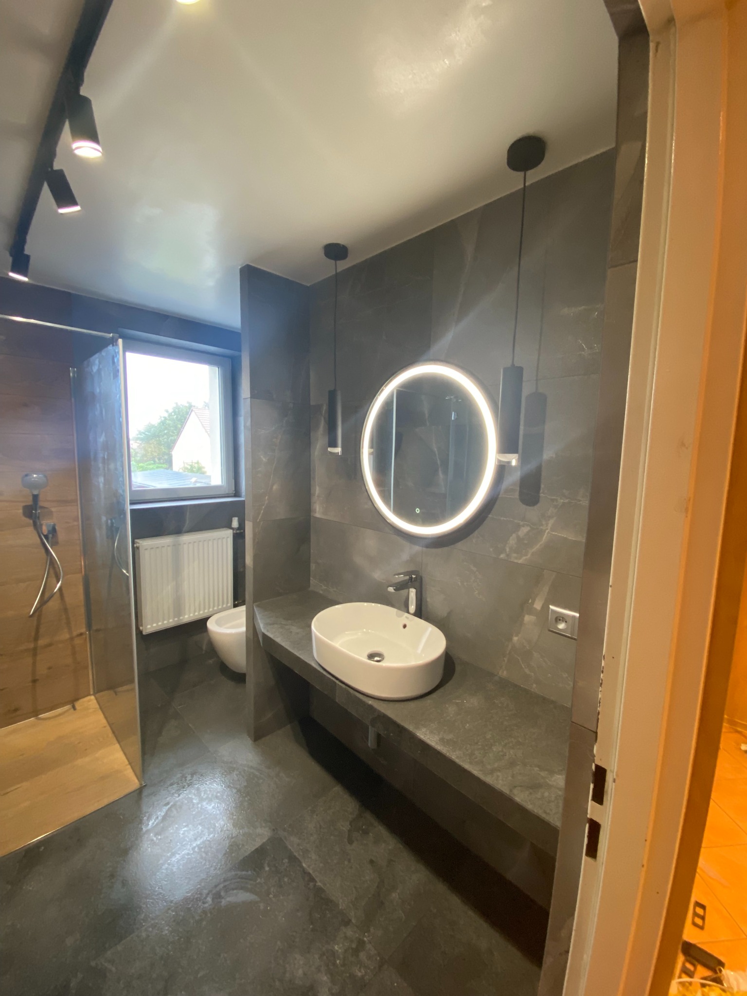 Nowoczesna łazienka z szarą armaturą, okrągłym lustrem z podświetleniem LED i kabiną prysznicową typu walk-in. Minimalistyczny design z akcentami drewna.