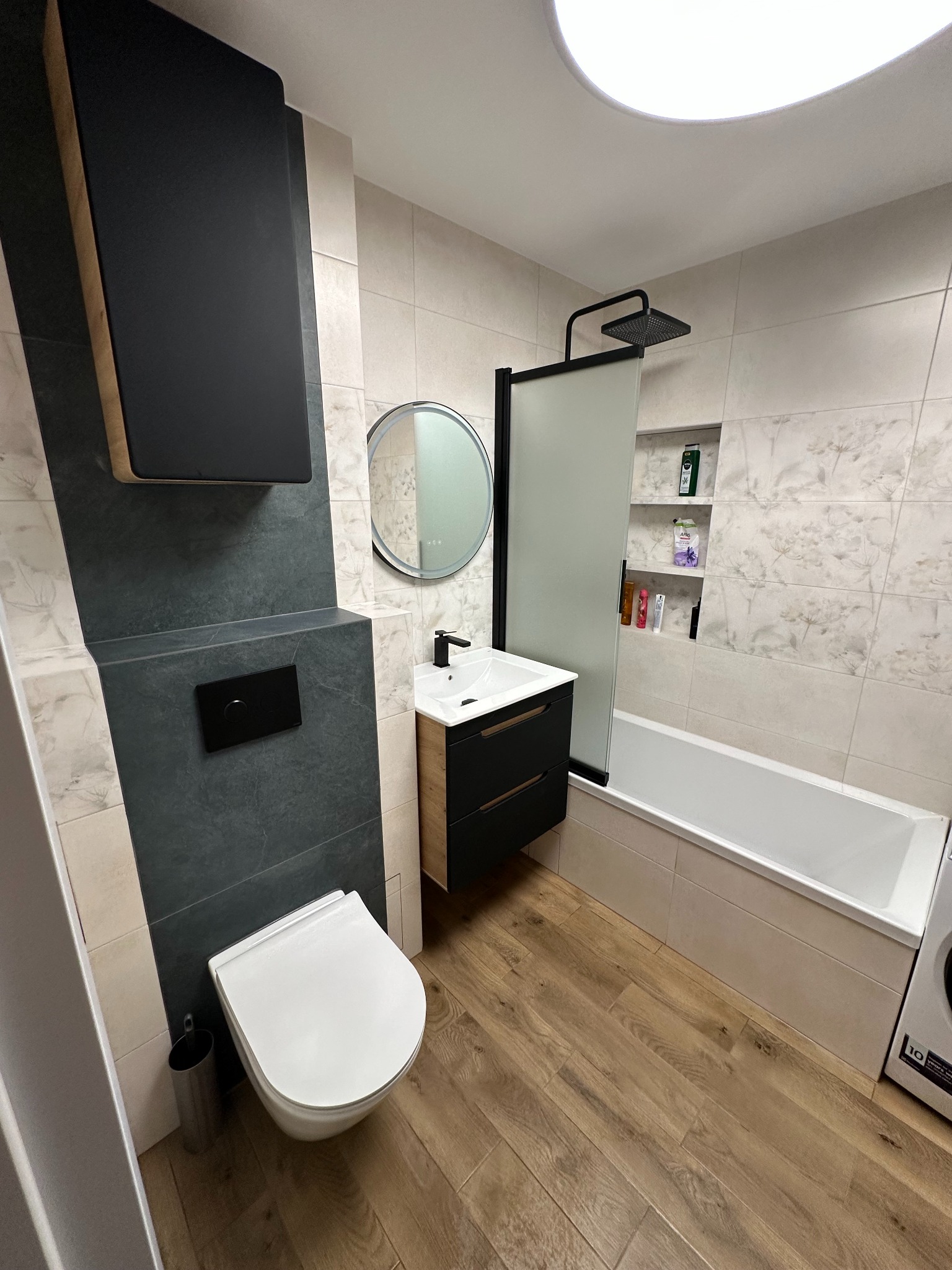 Nowoczesna łazienka z drewnianą podłogą, czarnymi akcentami i okrągłym lustrem. Uwagę przykuwa minimalistyczna armatura i subtelne oświetlenie.