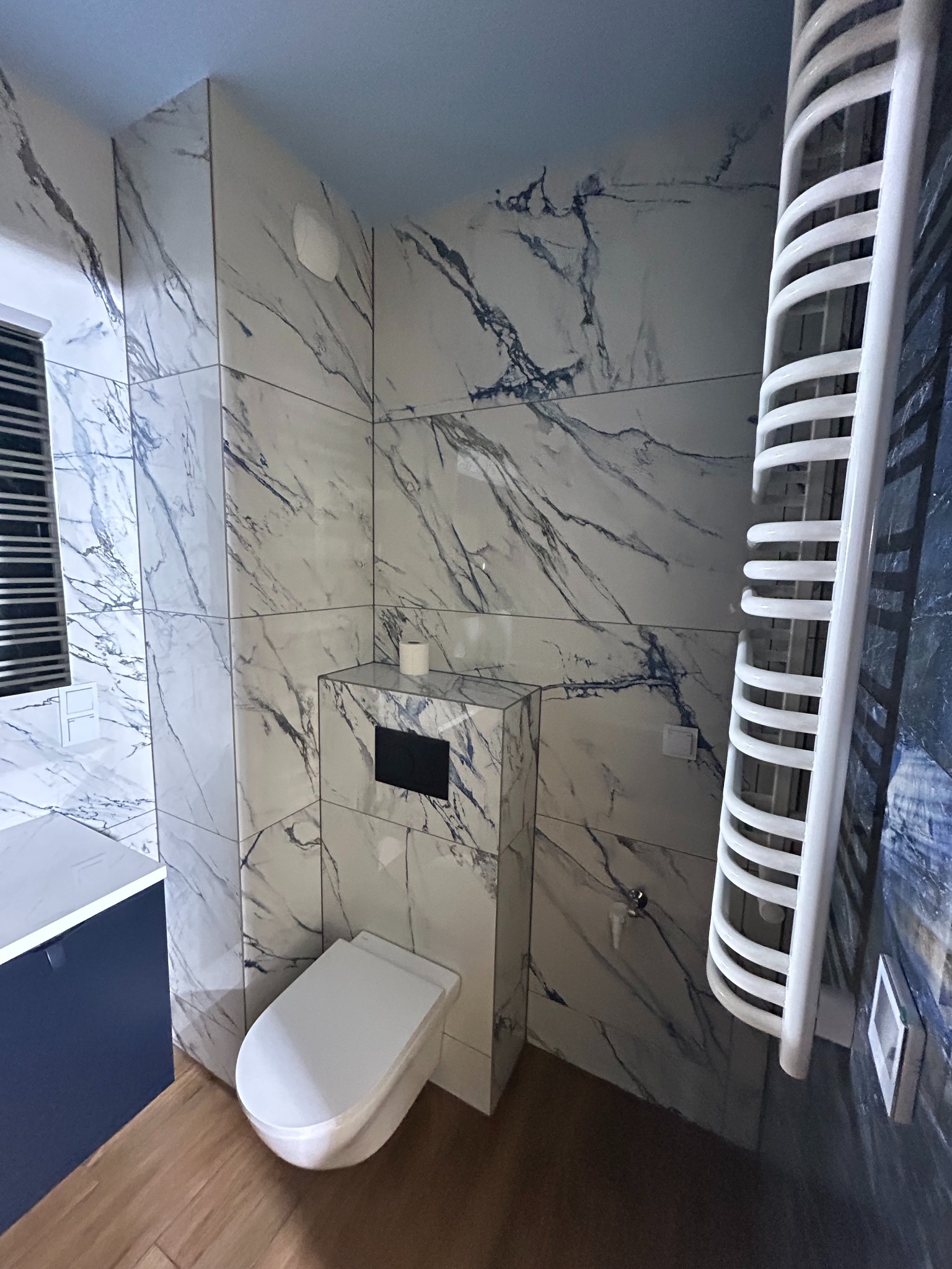 Nowoczesna łazienka z marmurowymi płytkami, podwieszaną toaletą i grzejnikiem drabinkowym. Uwagę przykuwa drewnopodobna podłoga i niebieska szafka umywalkowa.