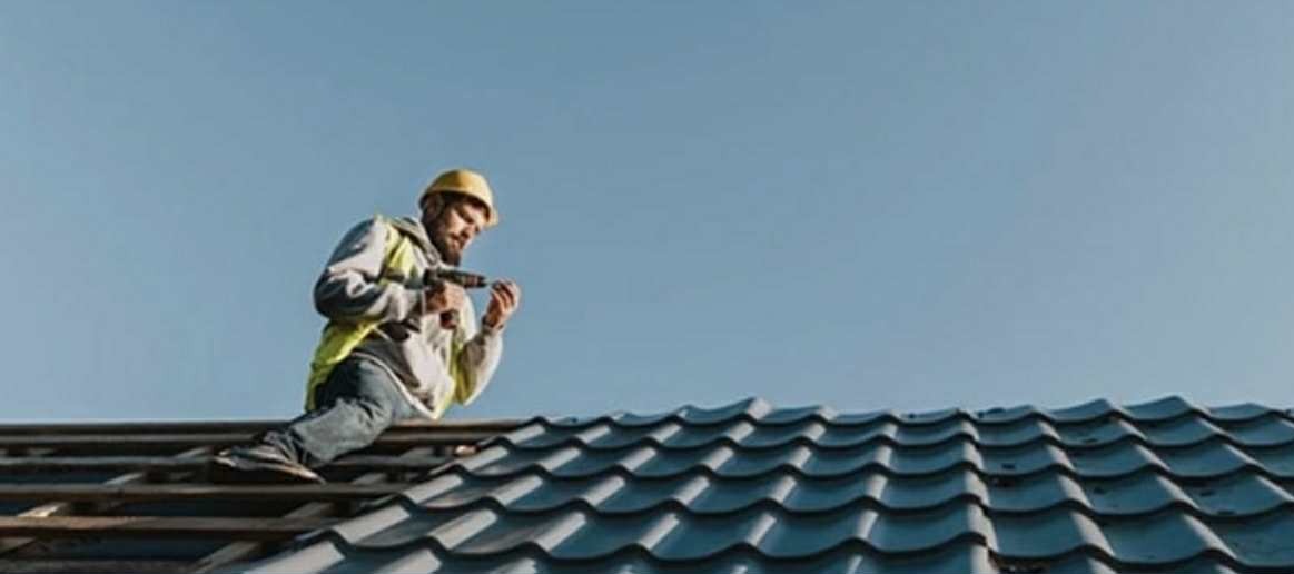 Dekarz w kasku montuje dachówkę na dachu domu. Widok z dołu na niebieskie niebo. Praca na wysokościach, narzędzia w dłoni.