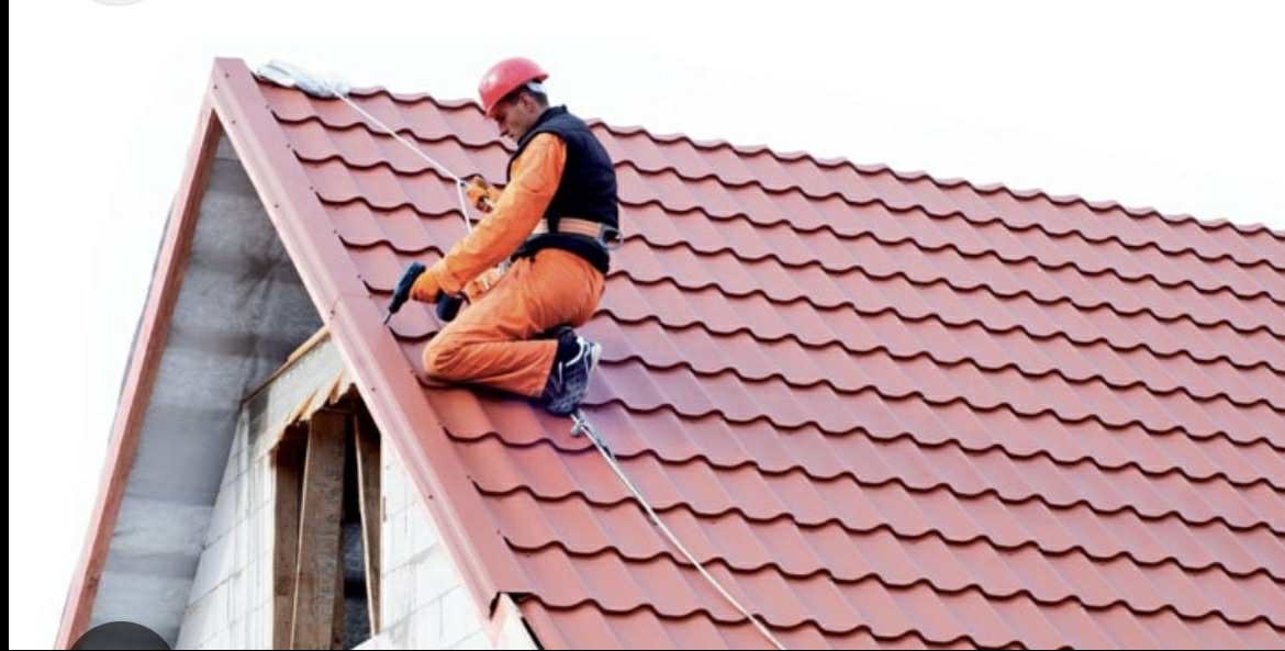 Pracownik w kasku, w trakcie montażu blachodachówki na skośnym dachu. Widoczne narzędzia i zabezpieczenia. Dach w odcieniach czerwieni, w tle fragment ściany budynku.