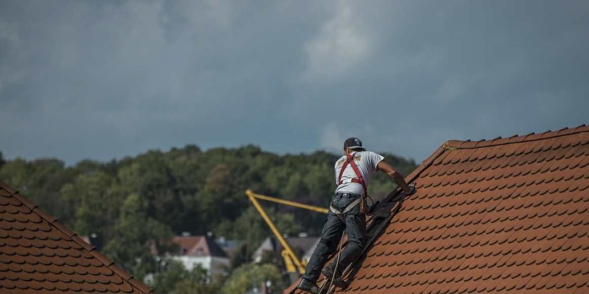 Dekarz w uprzęży na skośnym dachu z czerwonej dachówki, w tle zieleń drzew i fragmenty zabudowań. Praca na wysokości, zabezpieczenia.