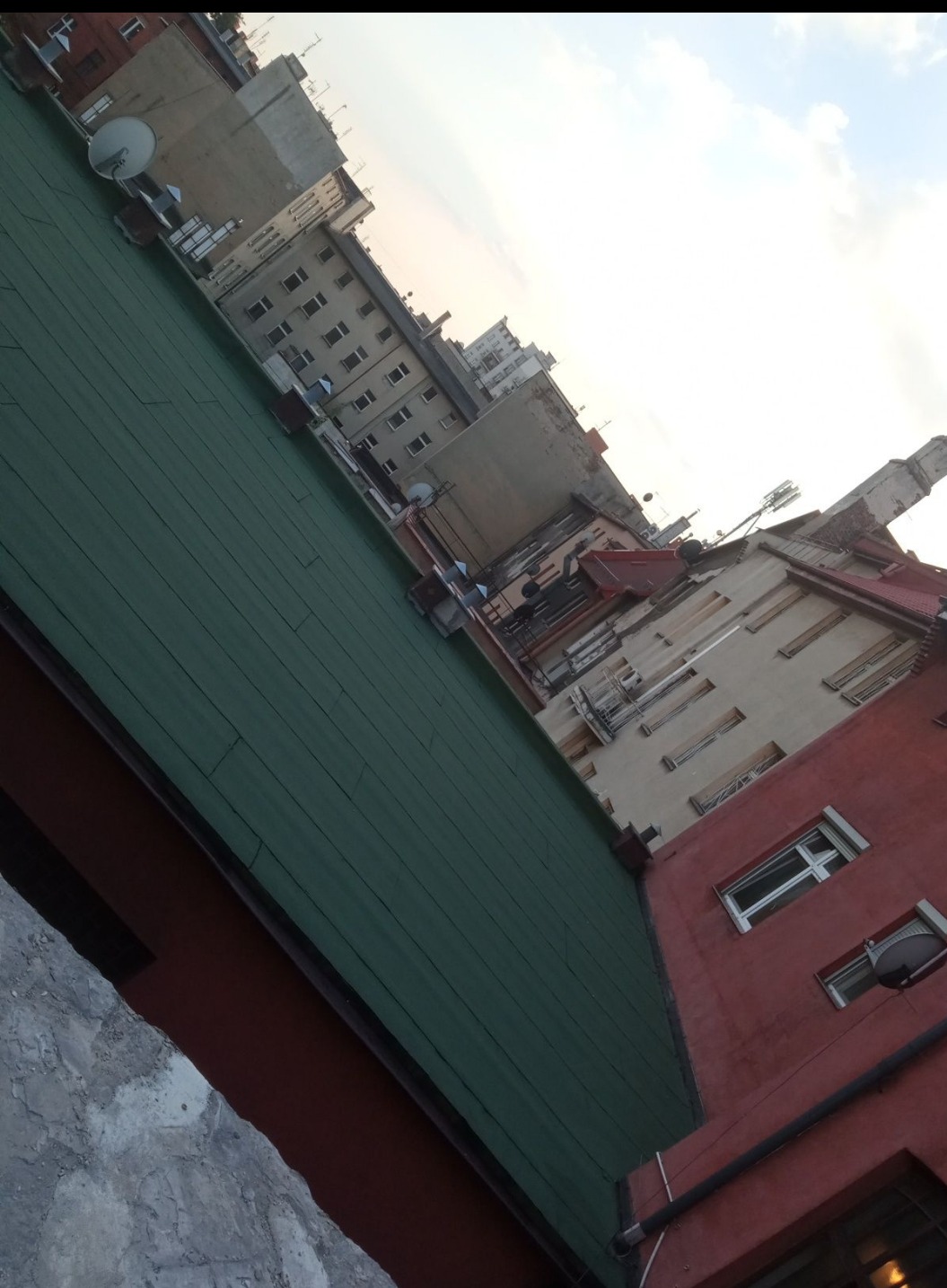 Zielony dach budynku z blachy na tle miejskiej zabudowy, ujęcie pod kątem. Widoczne budynki mieszkalne i fragment nieba.