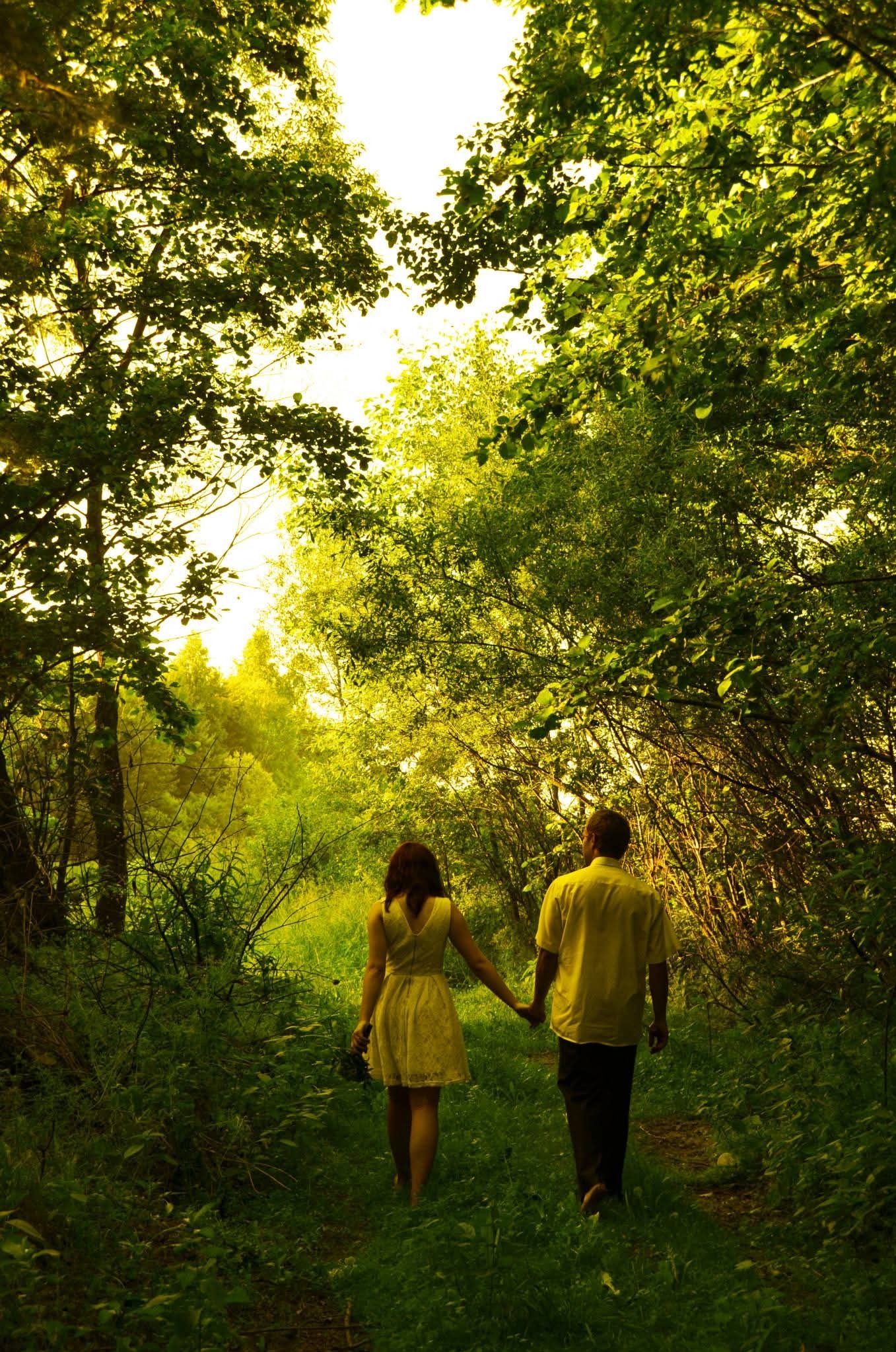 Para trzymająca się za ręce spaceruje leśną ścieżką w słoneczny dzień. Kobieta w żółtej sukience, mężczyzna w żółtej koszuli. Ujęcie z perspektywy osoby idącej za nimi.