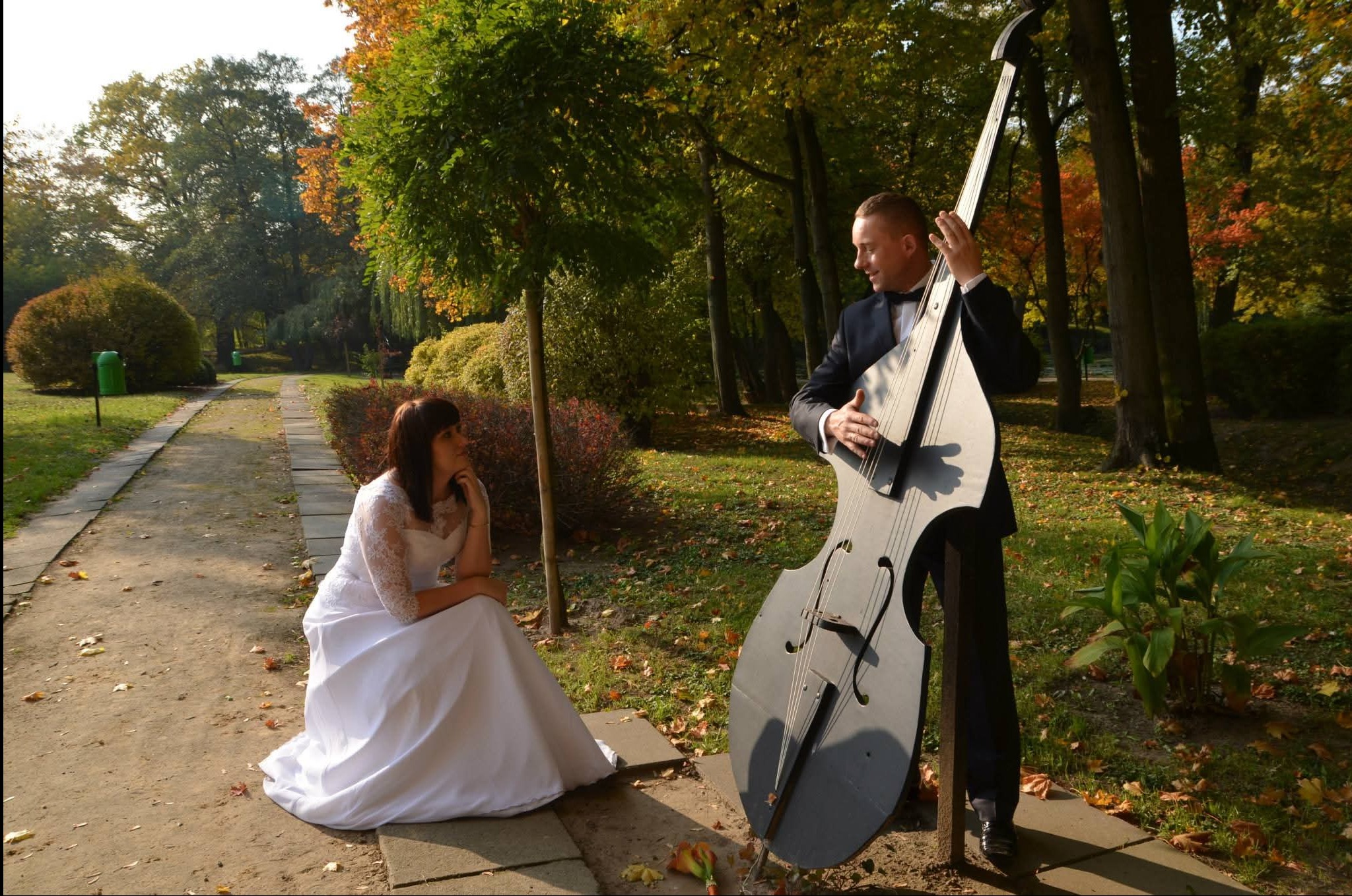 Jesienna sesja ślubna w parku: panna młoda w białej sukni siedzi na ścieżce, a obok stoi muzyk w garniturze z kontrabasem. Romantyczna atmosfera, ciepłe światło.
