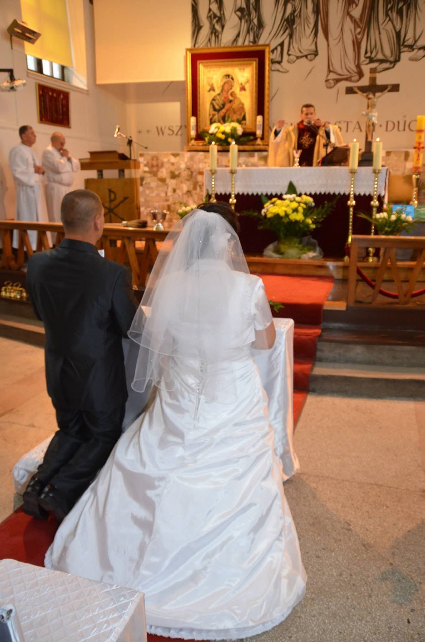 Para młoda klęczy podczas ceremonii ślubnej w kościele. Widok z tyłu na suknię panny młodej i garnitur pana młodego. W tle ołtarz, ksiądz i osoby w białych szatach.