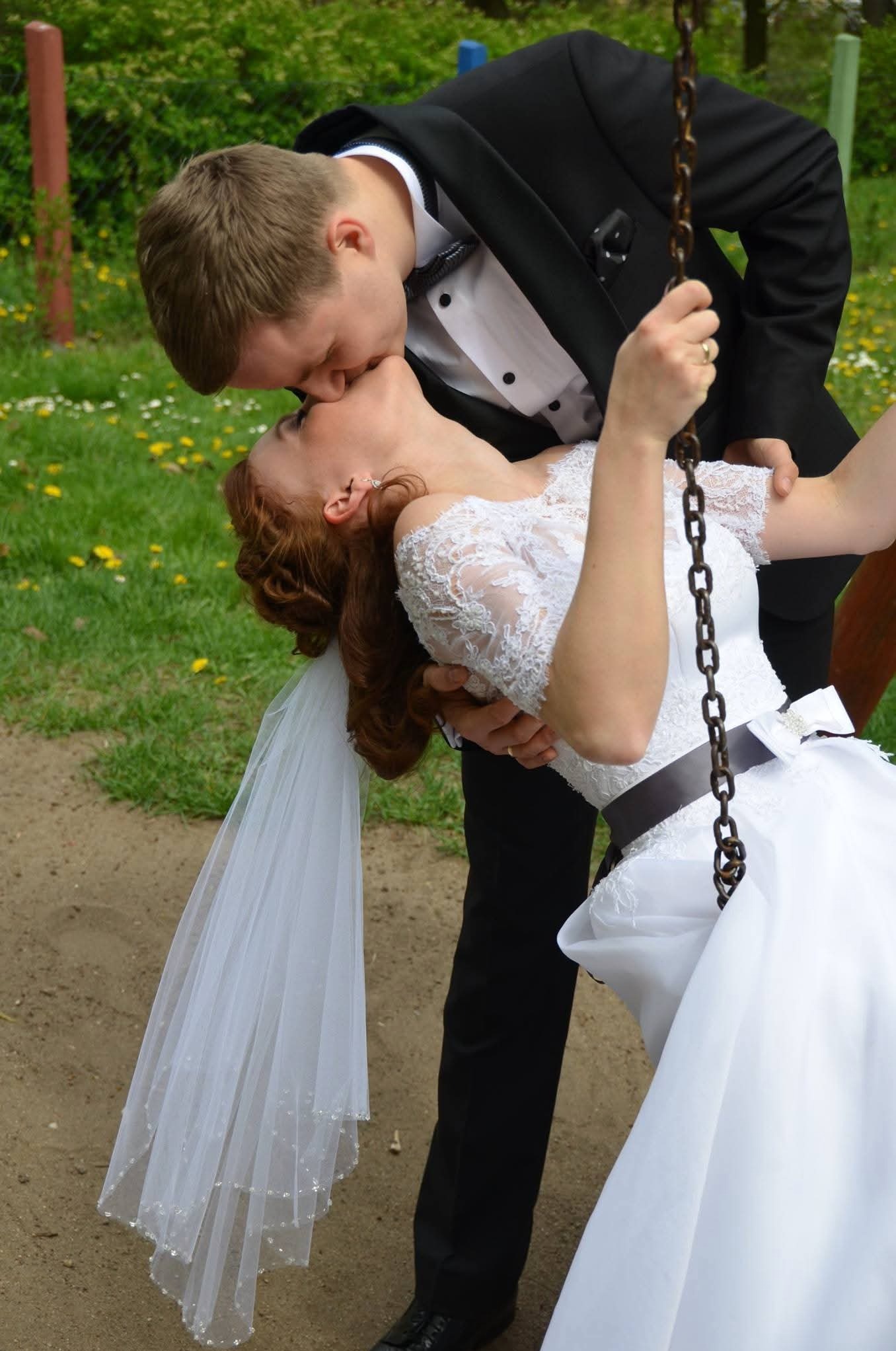 Para młoda w romantycznym uścisku na huśtawce w plenerze. Panna młoda w koronkowej sukni i welonie, pan młody w eleganckim smokingu. Ujęcie z bliska, podkreślające intymność chwili.