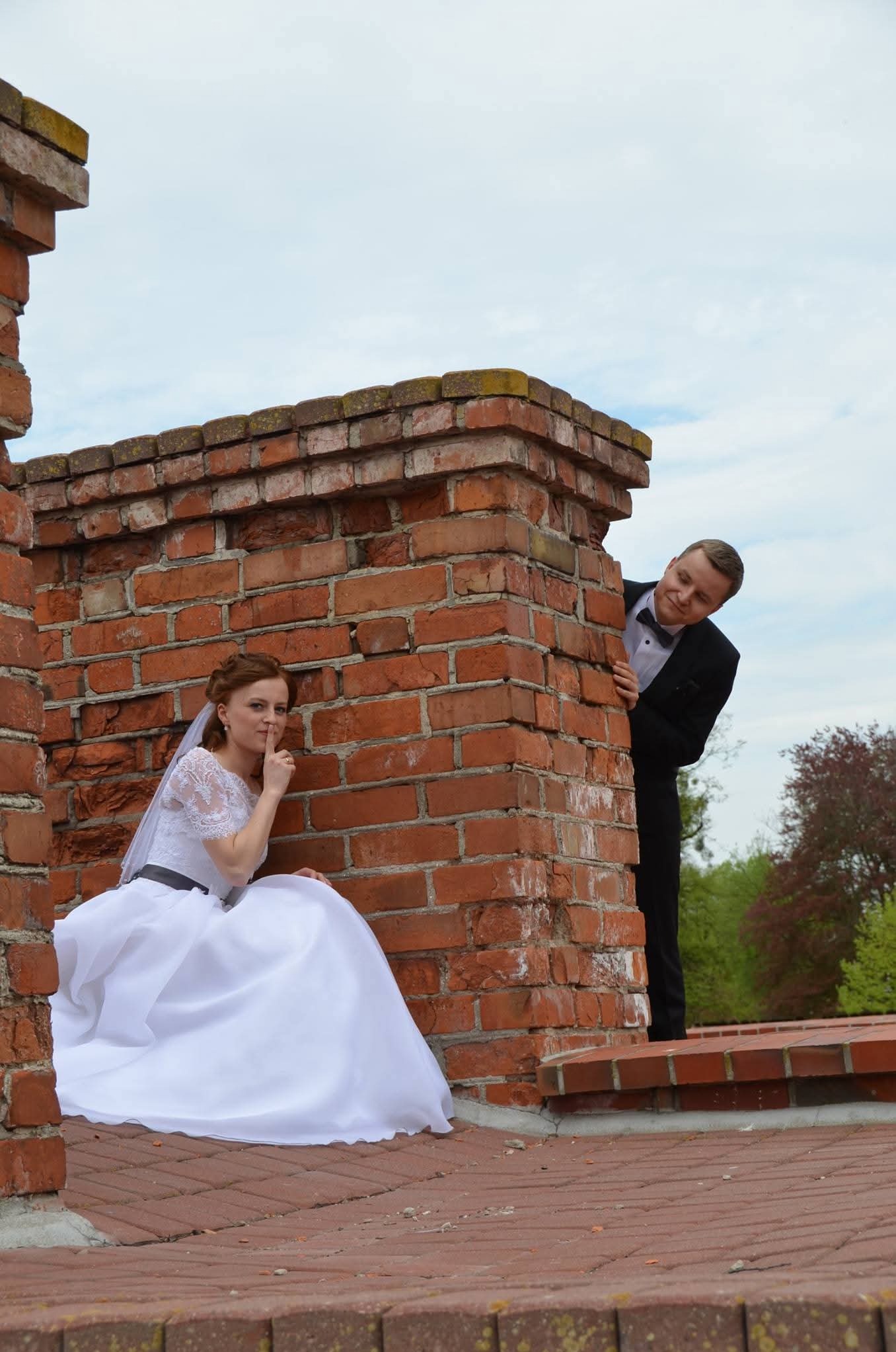 Para młoda w nietypowej scenerii: panna młoda siedzi za ceglanym murem, gestem ucisza, pan młody wygląda zza rogu. Romantyczna sesja w plenerze.