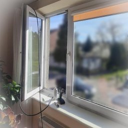 ASERWISOK Serwis I naprawa okien oraz drzwi PCV, ALUMINIOWE I Drewnianych - Białe okno PCV z otwartym skrzydłem, na parapecie wiertarka akumulatorowa, smar i narzędzia. Widok na zewnątrz, budynki w tle. Regulacja okna w trakcie.