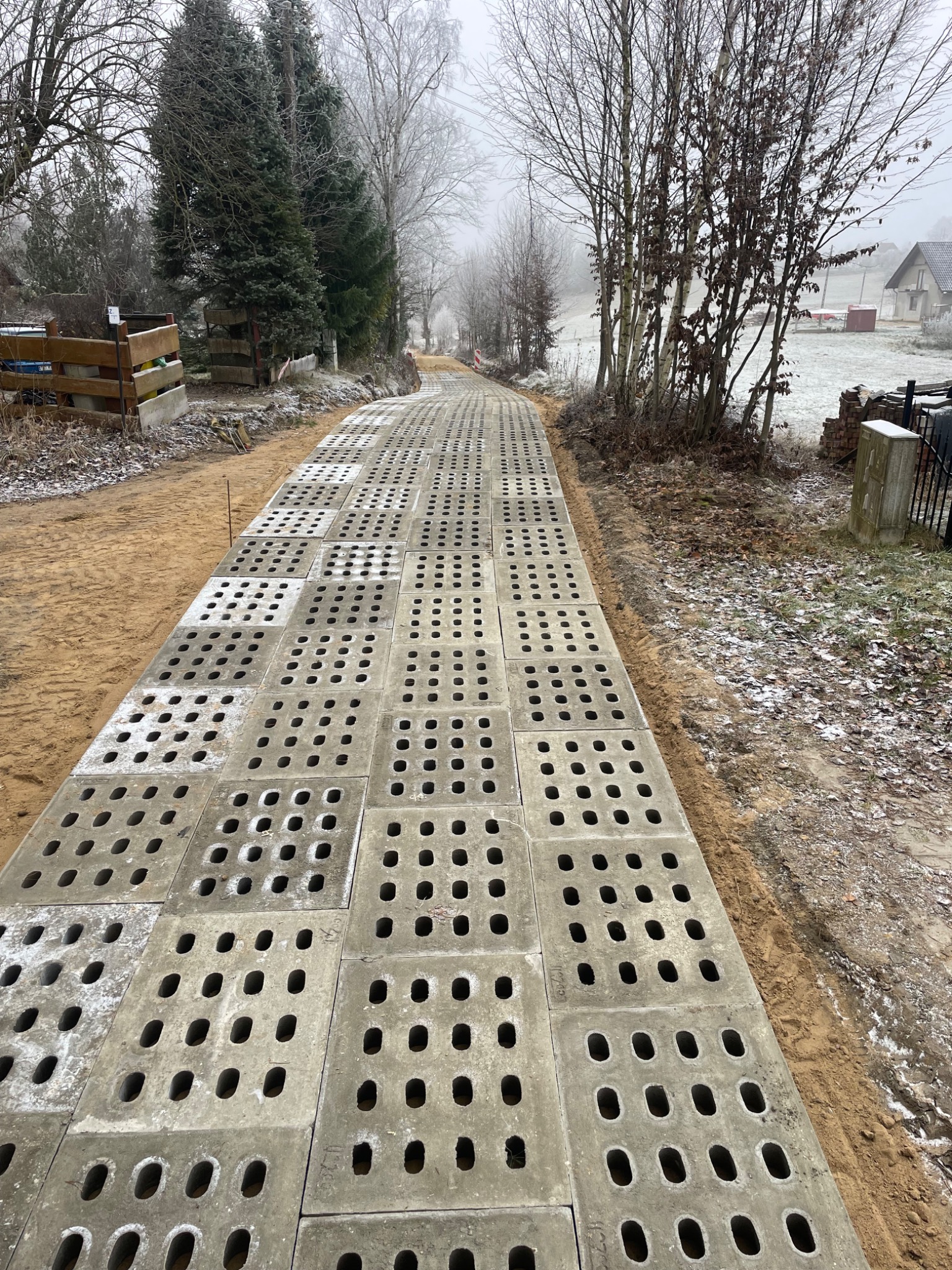 Ułożone betonowe płyty ażurowe na piaszczystym podłożu tworzące drogę dojazdową. Widok z perspektywy. Otoczenie: drzewa, trawa, budynek w tle. Pochmurny dzień.