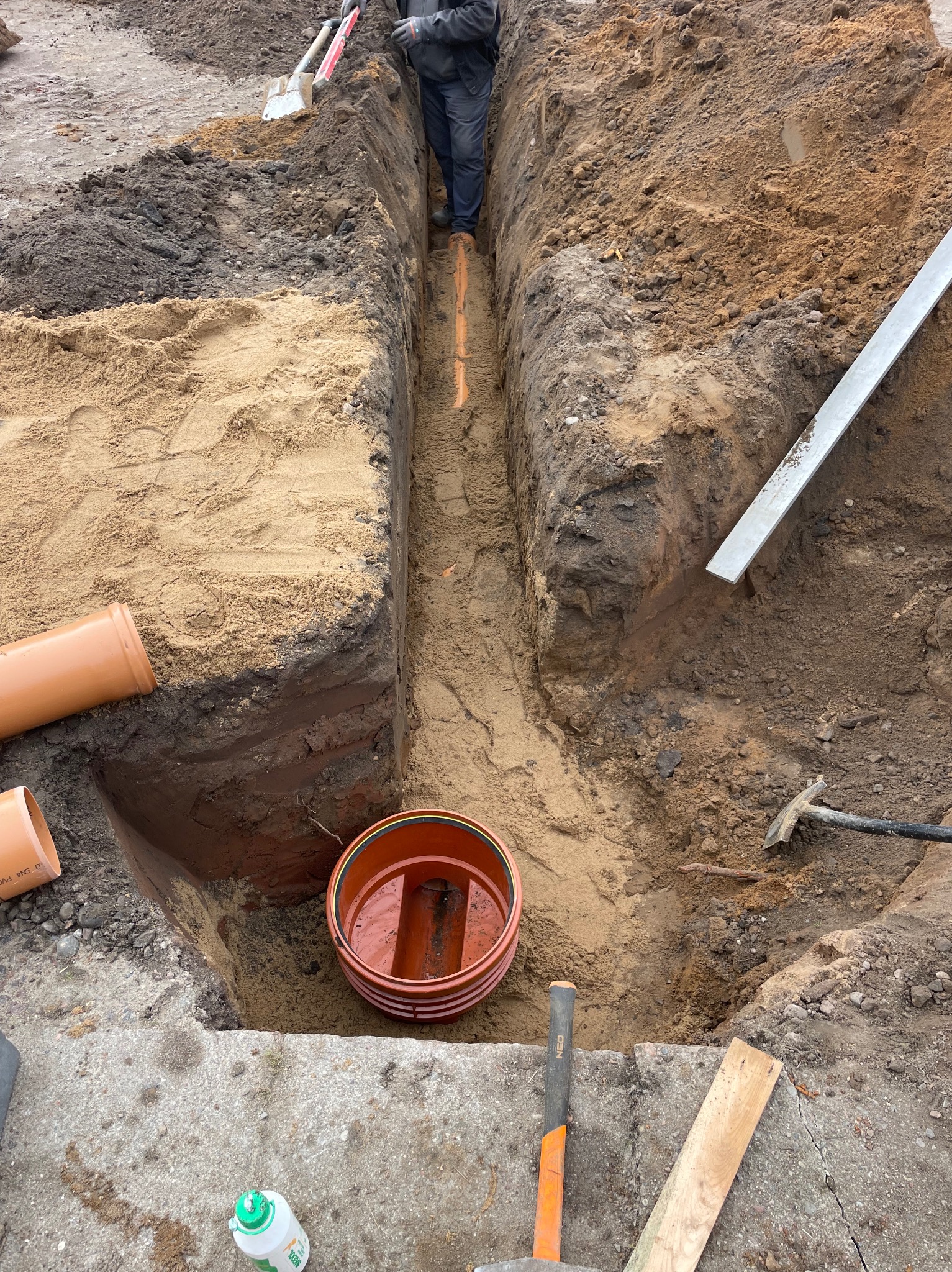 Wykop z pomarańczowymi rurami kanalizacyjnymi w ziemi, widoczne narzędzia i fragment osoby. Wykopane warstwy piasku i ziemi. Studzienka kanalizacyjna z wodą.