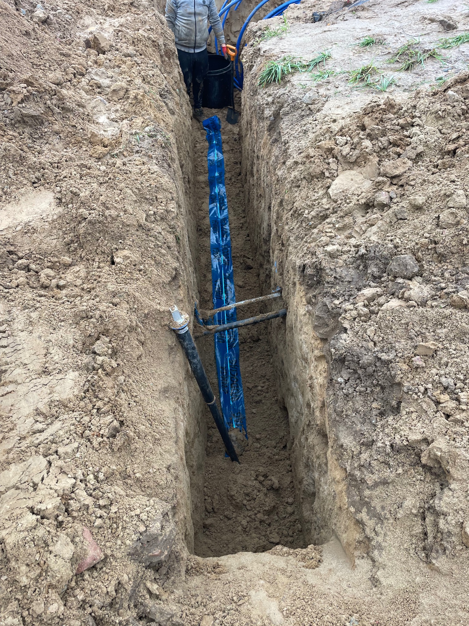 Wykopany wąski rów w ziemi z widoczną rurą wodociągową zabezpieczoną niebieską folią. W tle widoczna osoba w odzieży roboczej i fragment niebieskich rur.