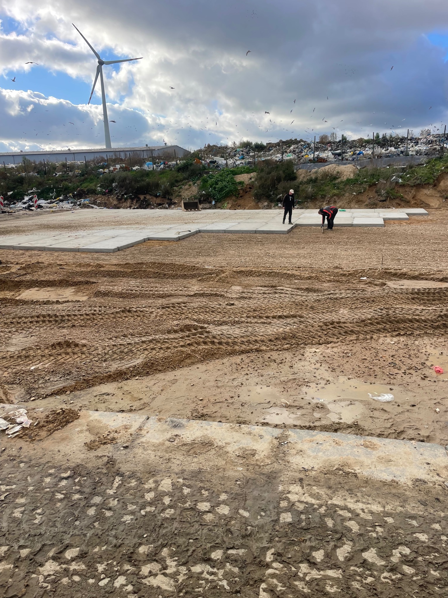 Układanie betonowych płyt na gruncie z widocznymi śladami opon, w tle składowisko odpadów i turbina wiatrowa. Dwóch pracowników przy pracy.