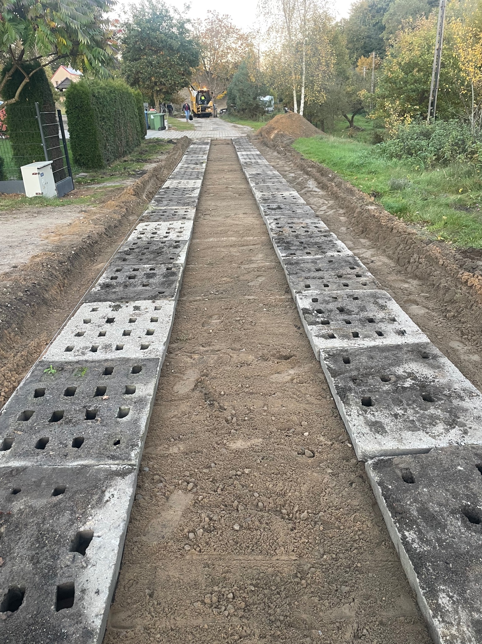 Długa, gruntowa droga z ułożonymi po bokach betonowymi płytami z otworami. W tle widoczna koparka i osoba. Otoczenie: drzewa i krzewy.