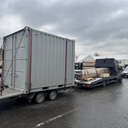 Auto Fox Karol Górski - Laweta z kontenerem i busem z ładunkiem na parkingu. Szare niebo w tle. Widoczne zabezpieczenia ładunku pasami. Transport towarów w pochmurny dzień.