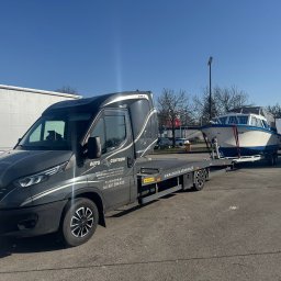 Auto Fox Karol Górski - Szara laweta Iveco z przyczepą, na której znajduje się biała łódź z niebieskim pasem. Widoczne logo firmy 'Auto Centrum' i dane kontaktowe. Błękitne niebo w tle.
