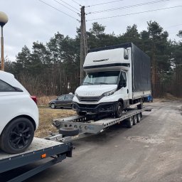 Auto Fox Karol Górski - Laweta z białym Iveco i Oplem Corsa na platformie, na szarym asfalcie, w tle las i słup oświetleniowy. Widok z boku, pochmurny dzień.