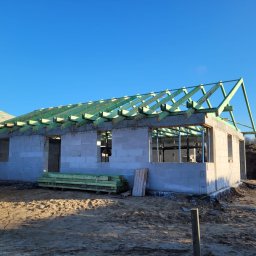 USŁUGI OGÓLNOBUDOWLANE MATEUSZ ZAKRZEWSKI - Budowa domu z betonu komórkowego w stanie surowym otwartym, widoczne ściany, więźba dachowa zaimpregnowana na zielono, paleta bloczków H+H. Słoneczny dzień.