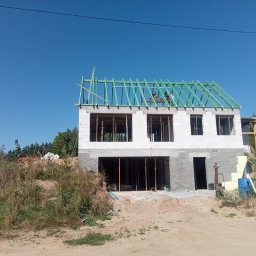 USŁUGI OGÓLNOBUDOWLANE MATEUSZ ZAKRZEWSKI - Dwukondygnacyjny dom w trakcie budowy: białe ściany z betonu komórkowego, zielona konstrukcja dachu, widoczne zbrojenia okien. Prace budowlane w toku, słoneczny dzień.