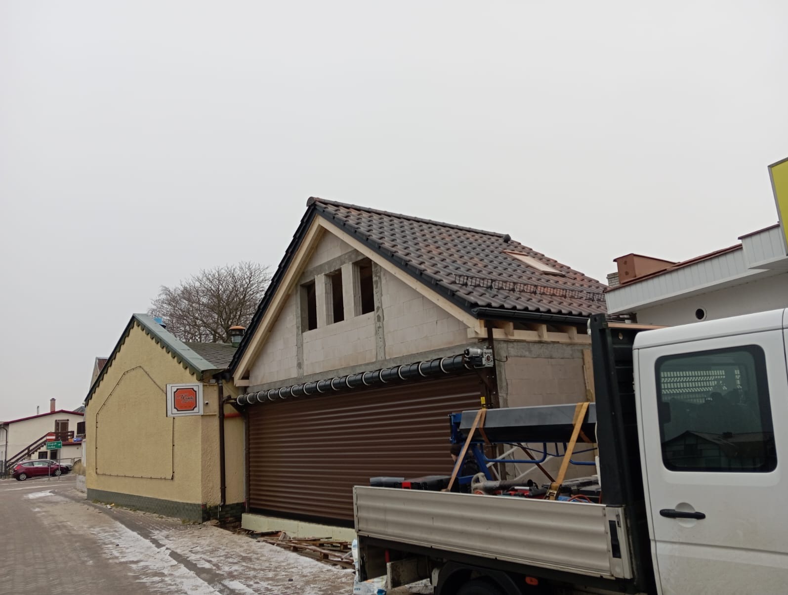 Rozbudowa domu z keramzytu: widok na dobudowaną część z oknami, brązową bramą rolowaną i fragmentem dachu z dachówką. Na pierwszym planie fragment samochodu dostawczego.