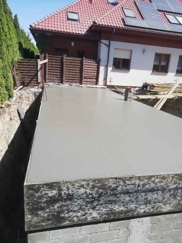 Zalany fundament pod budowę domu jednorodzinnego z widoczną konstrukcją szalunkową i wystającymi prętami zbrojeniowymi. W tle dom z panelami słonecznymi na dachu.