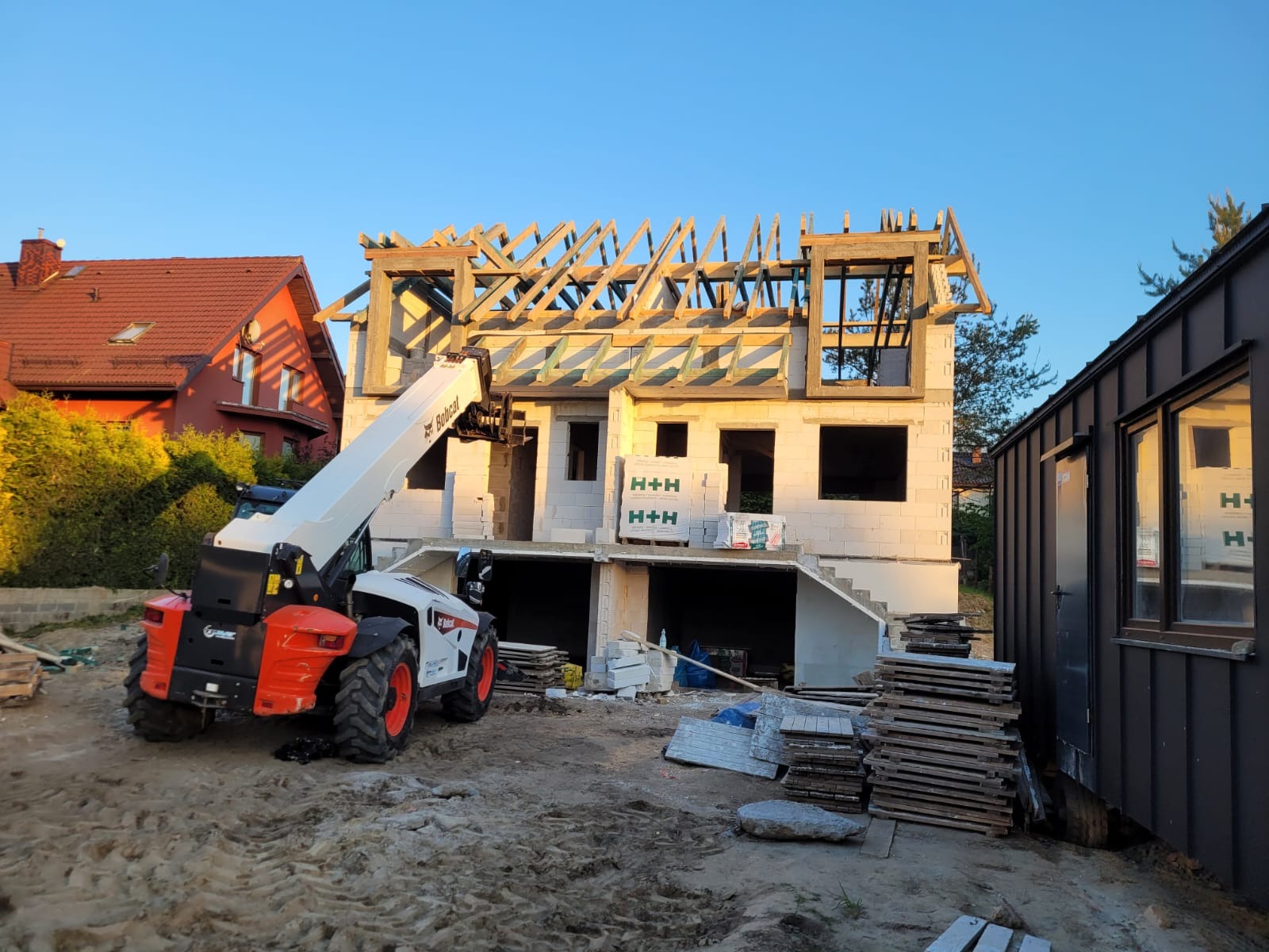 Budowa domu z białego pustaka H+H, widoczny dźwig Bobcat, drewniana konstrukcja dachu w trakcie montażu, w tle dom z czerwoną dachówką i budynek z ciemną elewacją.