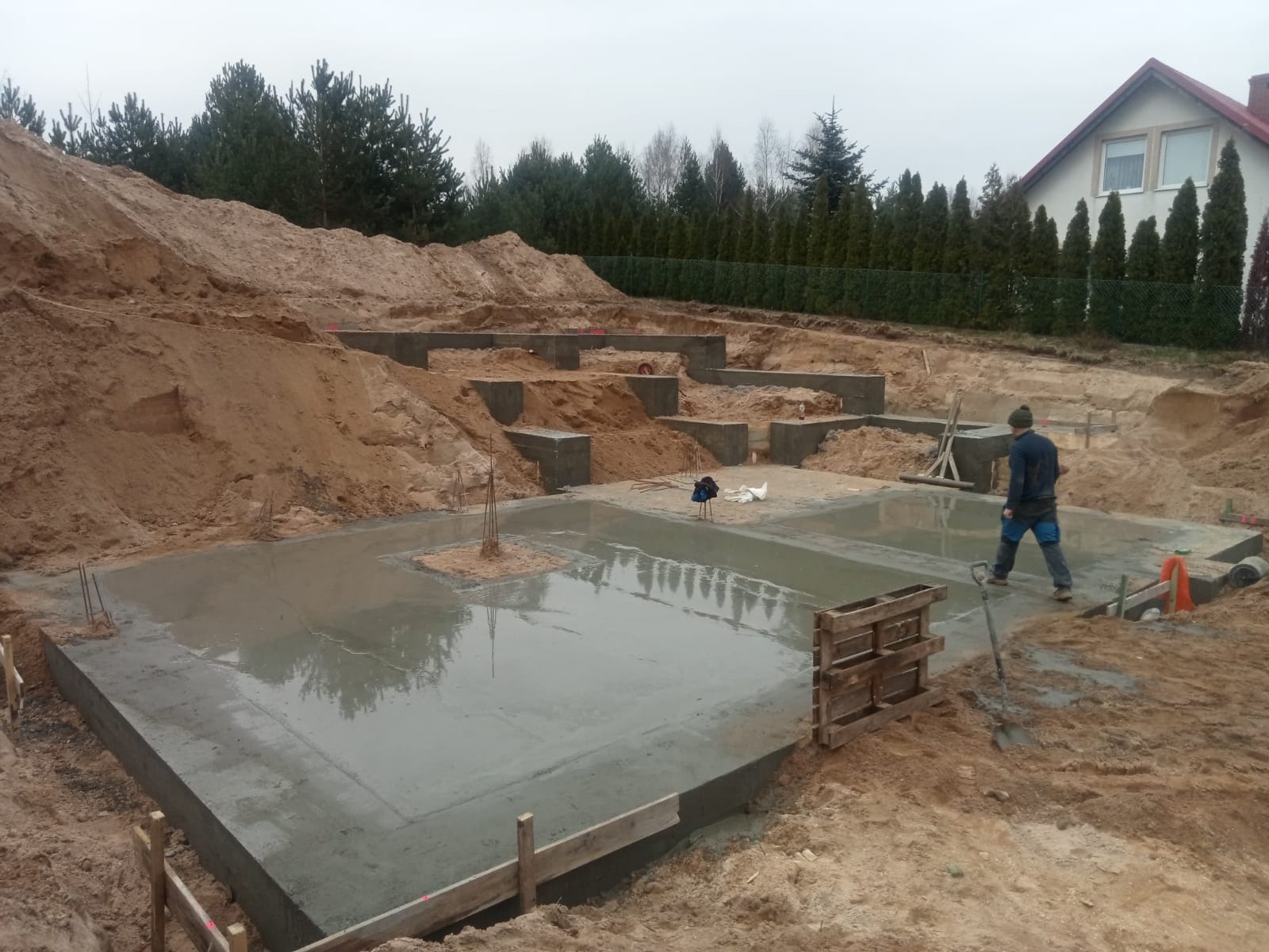 Zalany fundament domu jednorodzinnego, widoczne zbrojenia i szalunki. W tle wykopana ziemia i zieleń. Pracownik w roboczym ubraniu na terenie budowy.
